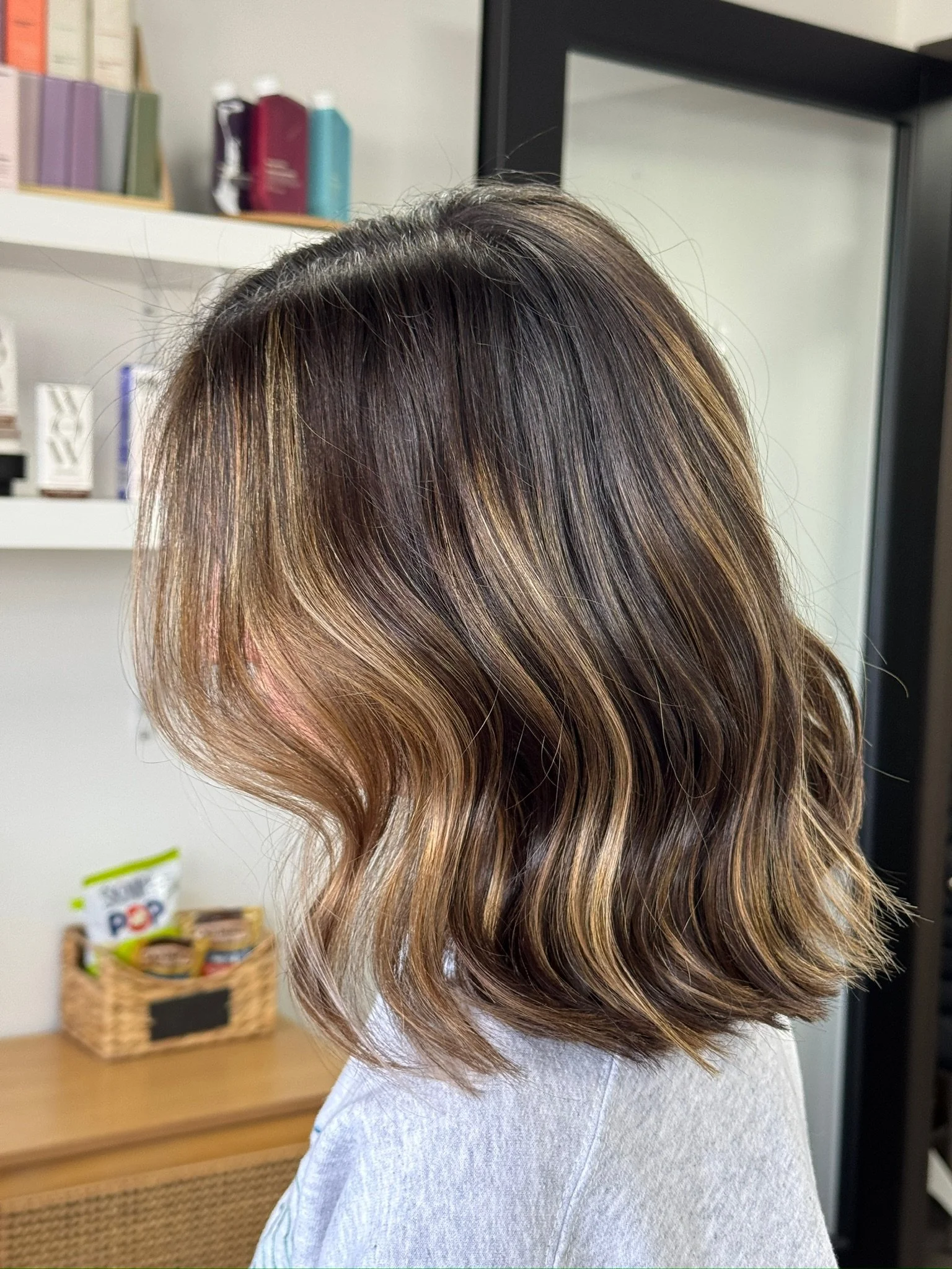 Close-up of a person's hair styled in loose wavy layers with blonde highlights, seen from the back in a salon or home setting.