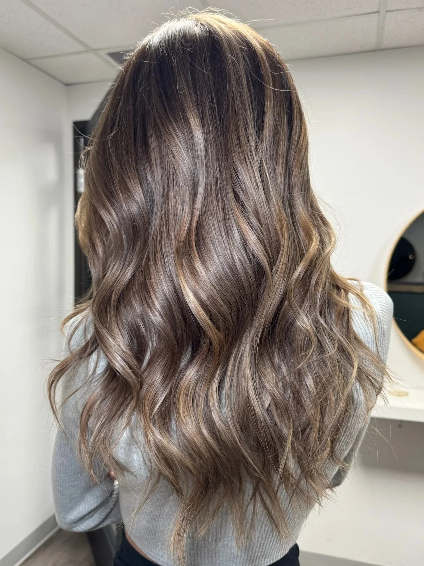 Back view of a woman with long, wavy hair that has balayage highlights in shades of blonde and light brown.