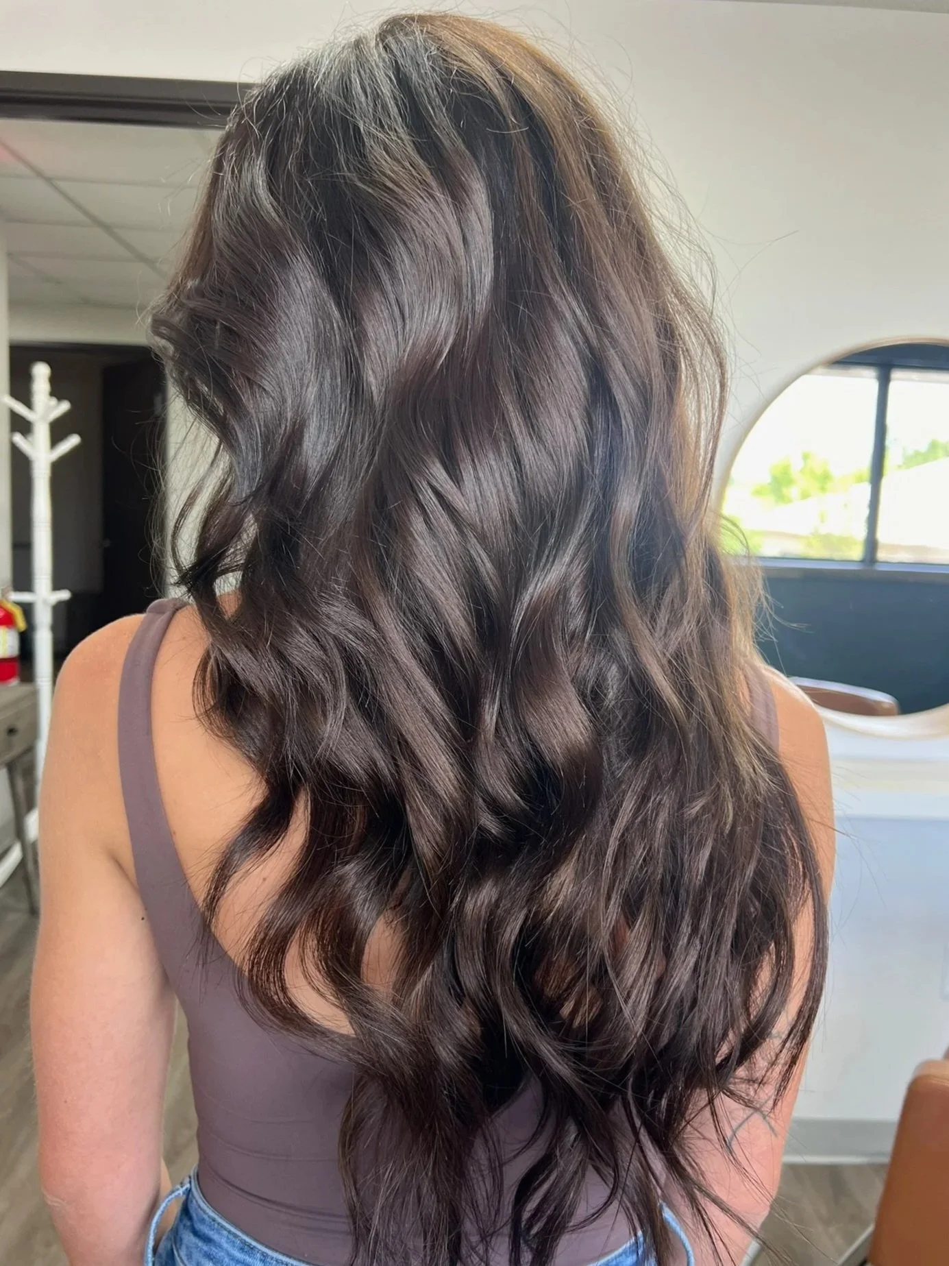 Long, wavy, dark brown hair worn loose over a person's shoulders with sunlight highlighting the hair. The person is wearing a sleeveless, purple top and sitting in what appears to be a salon or indoor setting.