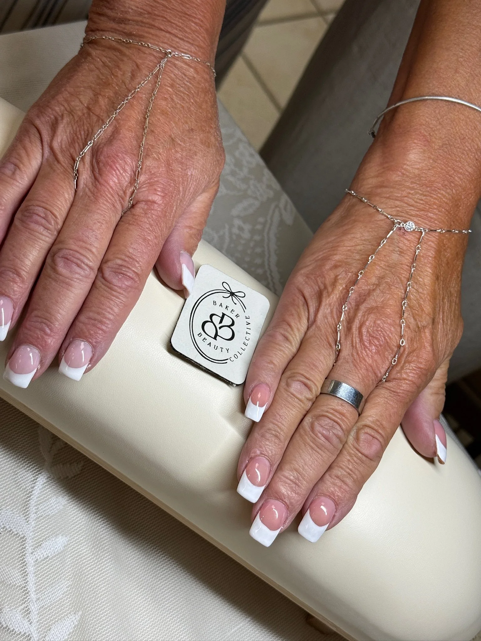 Close-up of a person's hands with manicured nails, wearing jewelry including rings and a bracelet. The hands rest on a light-colored surface with a bakery and beauty collection tag visible.