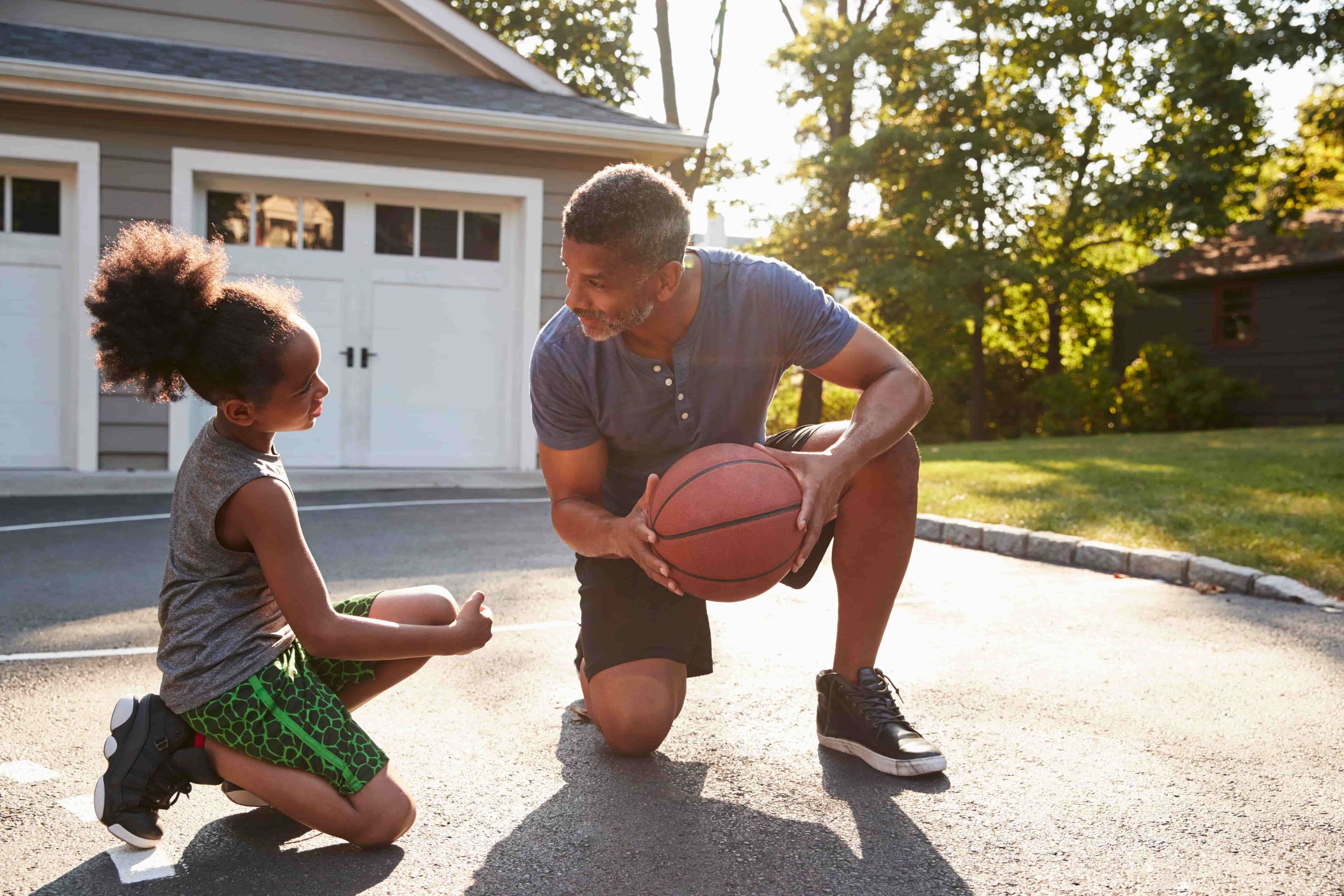 A young girl and an older man play basketball outdoors in a driveway near a garage, with trees and sunlight in the background.