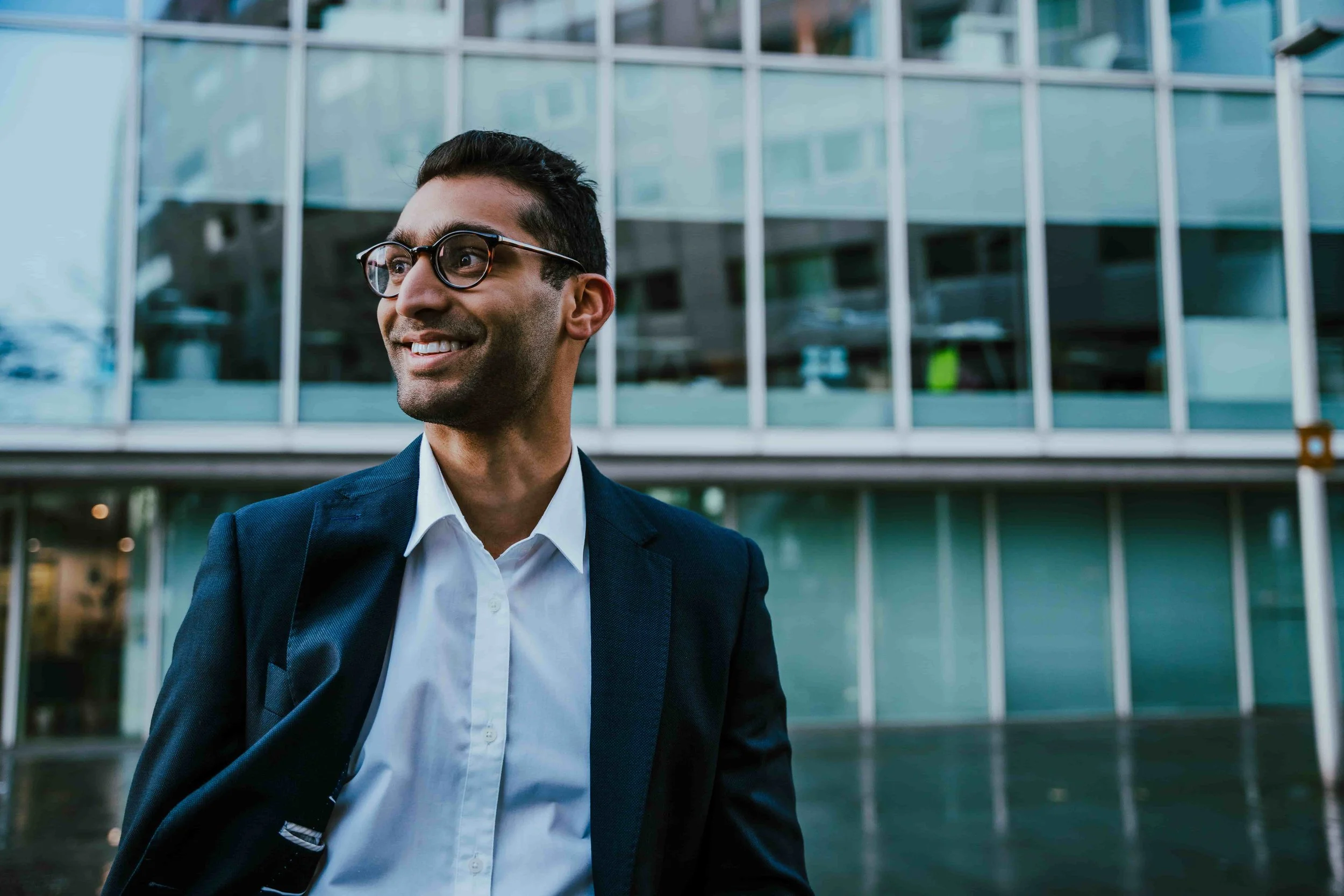 A man wearing glasses and a navy suit smiling outdoors in front of a modern glass building.