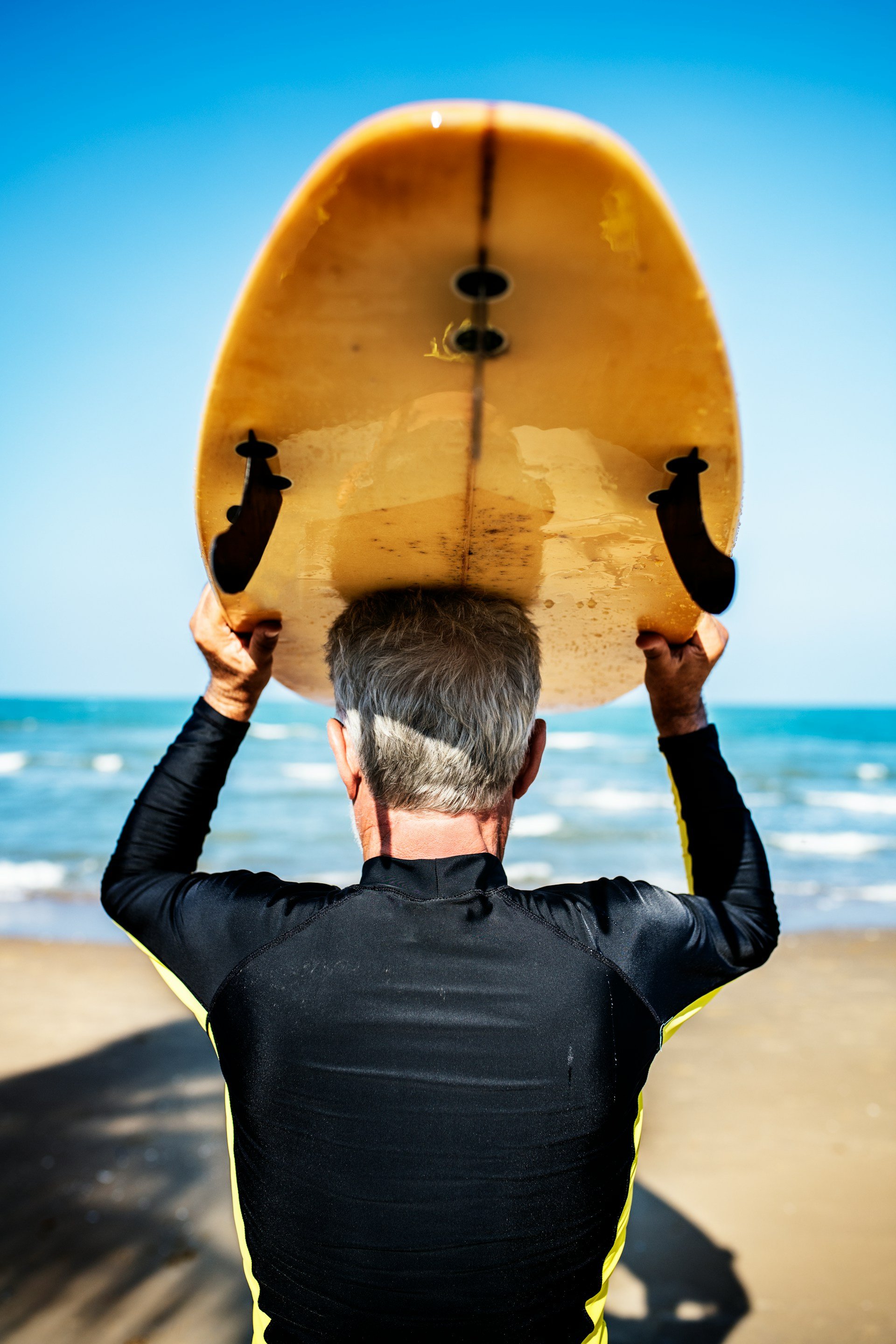 Person holding a yellow surfboard above their head on a sandy beach with the ocean and sky in the background.