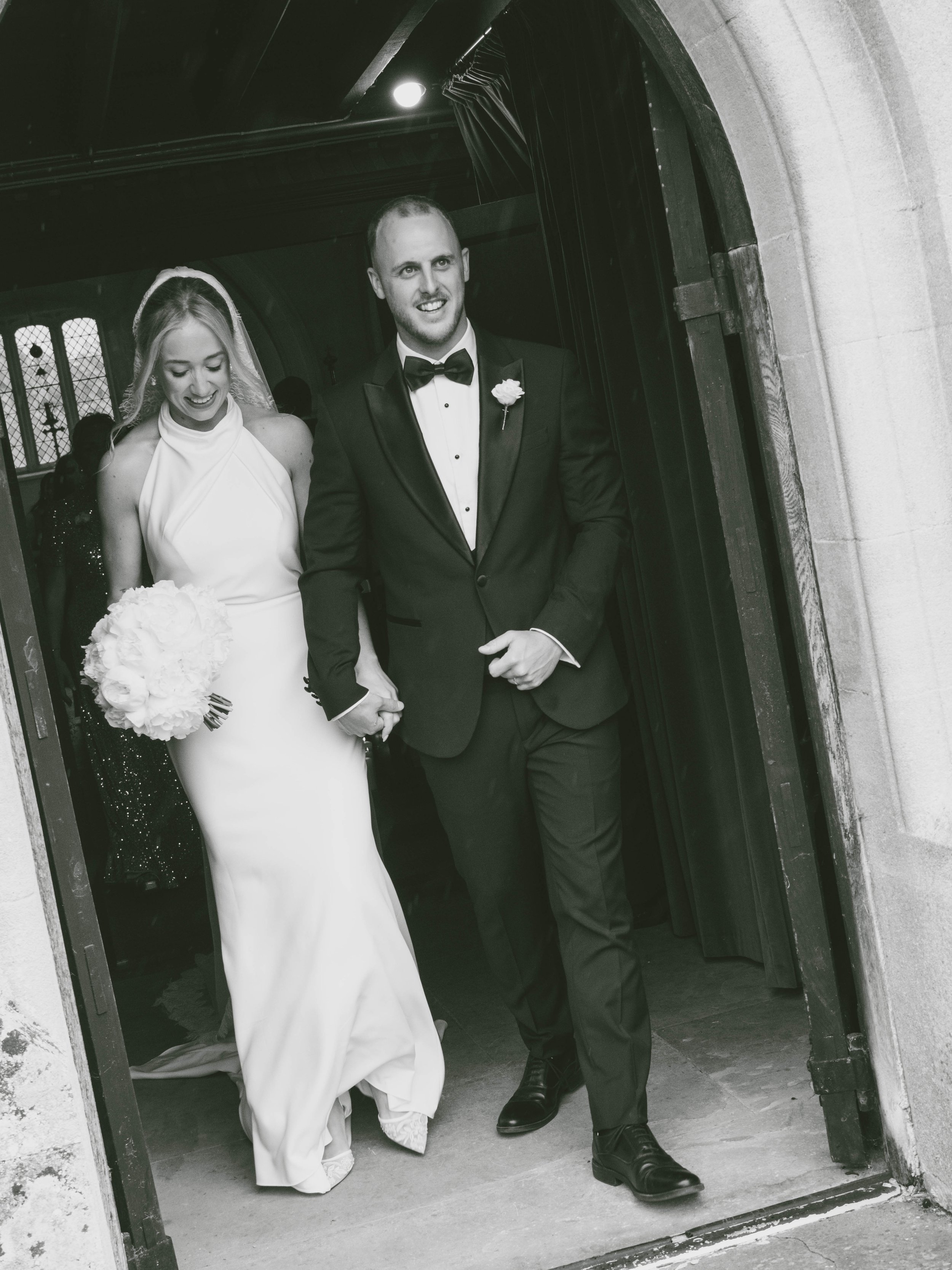 A black and white photo of a bride and groom walking through a doorway with stone framing, holding hands and smiling, dressed in wedding attire.