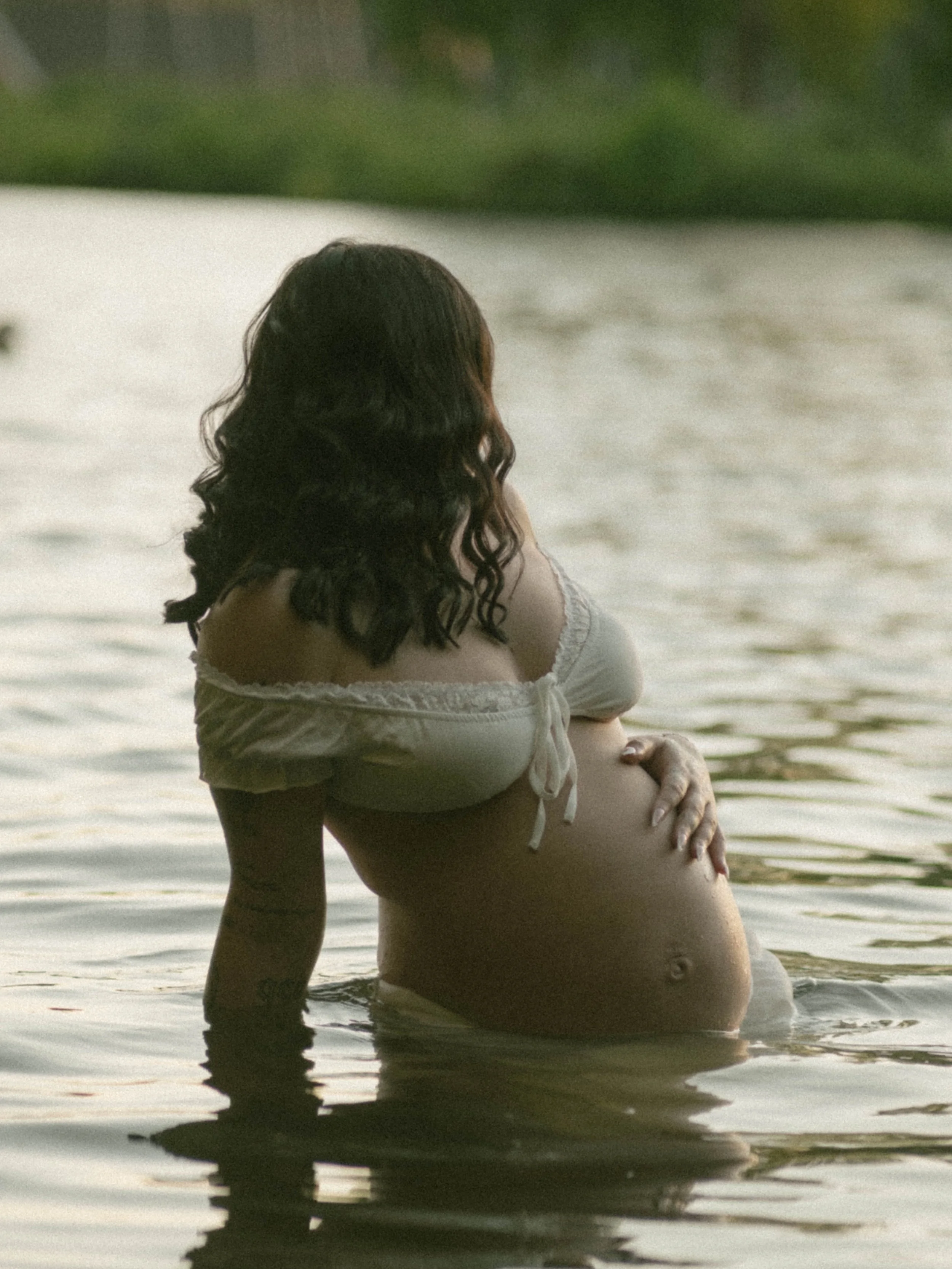 A woman with dark, wavy hair is sitting in the water at a beach or lake, facing away from the camera, with a green landscape in the background.