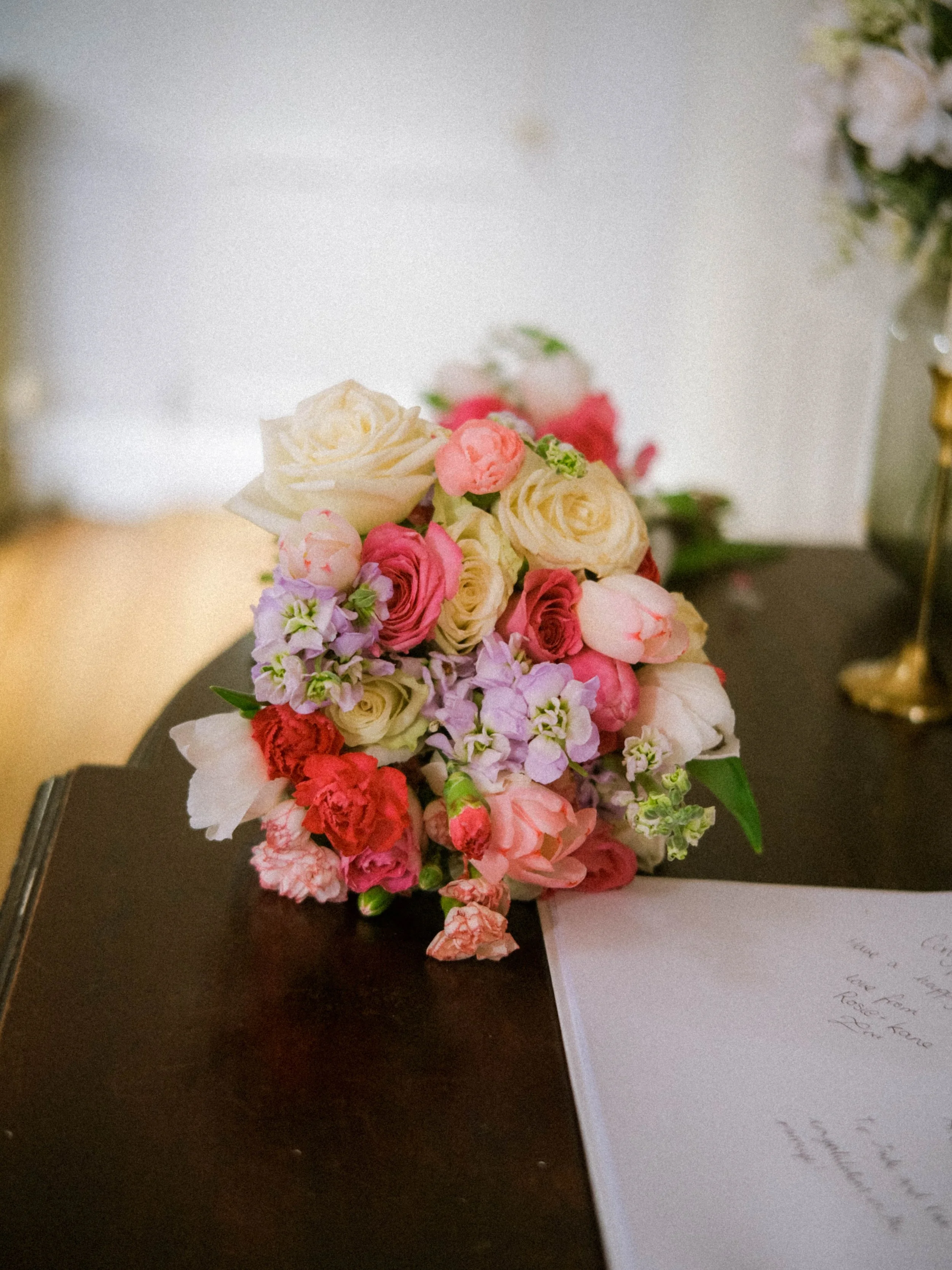 Colorful bouquet of flowers including roses and other blooms on a dark wooden surface with a handwritten letter beside it.