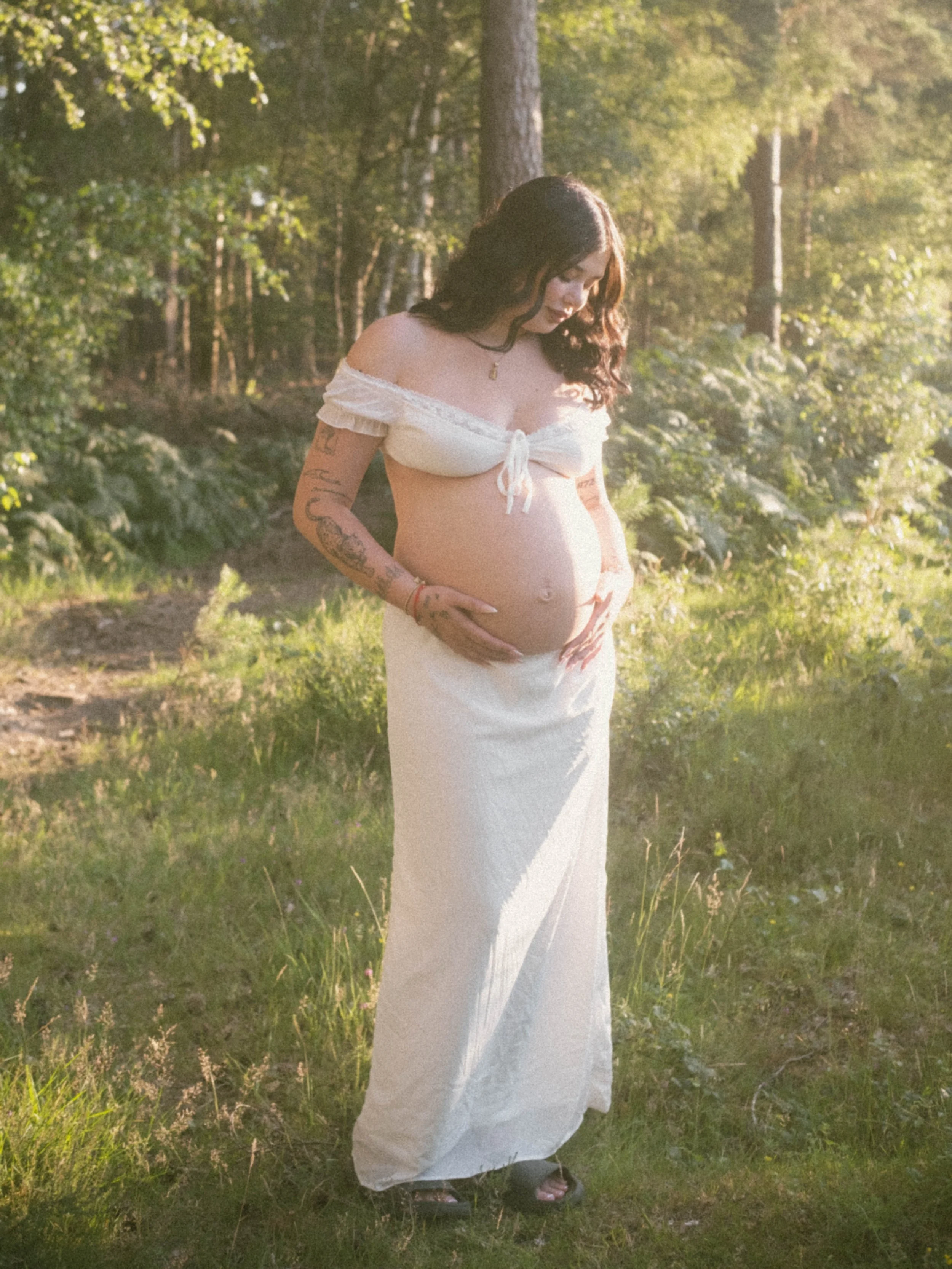 A pregnant woman with dark curly hair wearing a white off-the-shoulder crop top and a long white skirt stands outdoors in a wooded area during sunlight, gently cradling her belly.