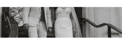 A bride and groom standing on the steps of a building, holding hands, with the bride wearing a lace dress and the groom in a suit.