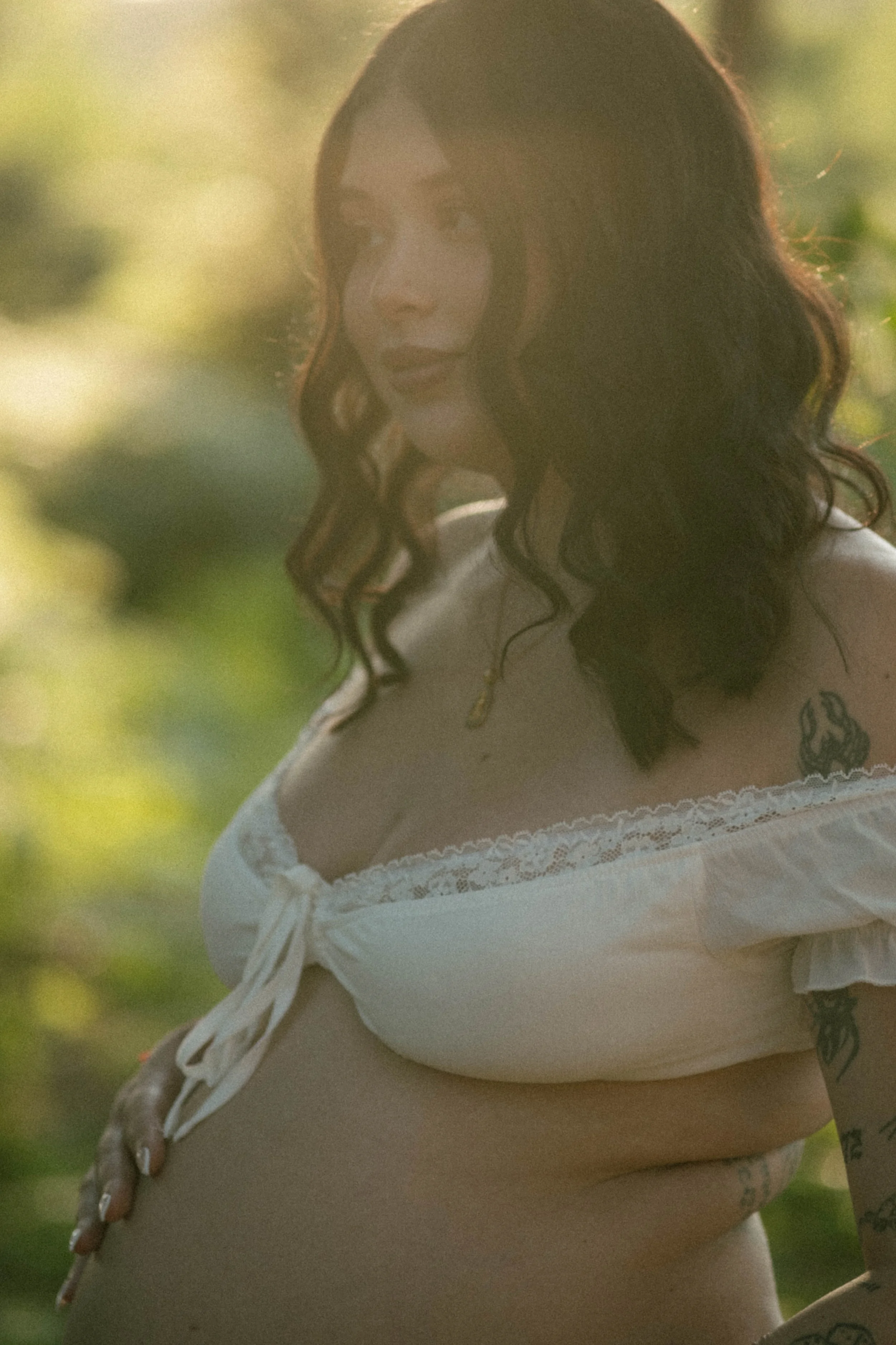 A woman with long, curly dark hair, wearing a white lace blouse with tied sleeves, stands outdoors with a blurred green background, and has tattoos on her arm and shoulder.