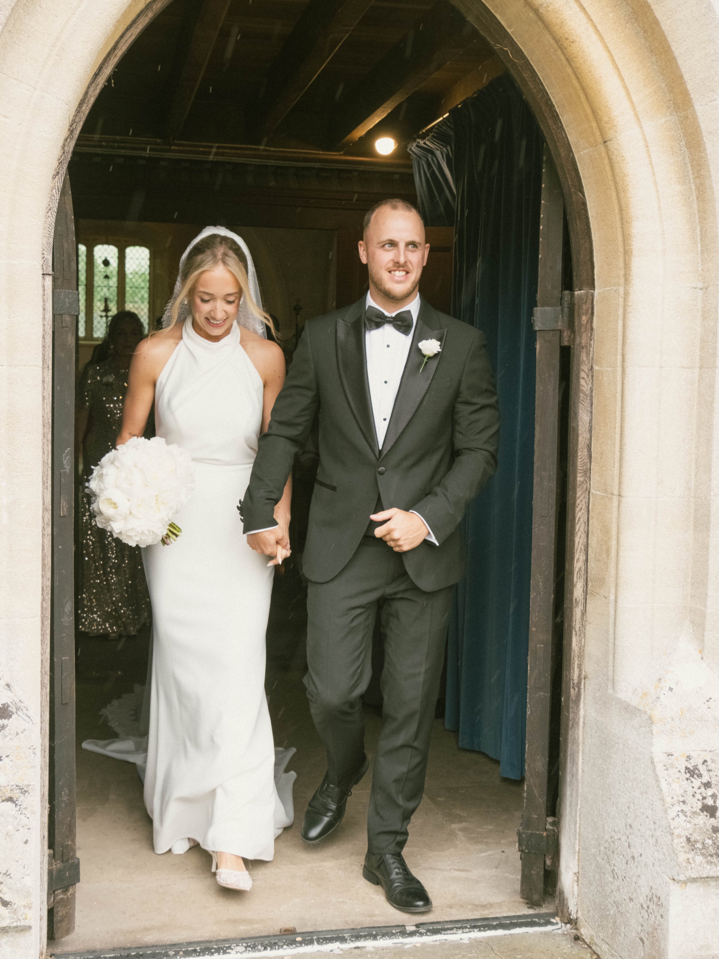 A newlywed couple walking hand in hand through a church doorway during their wedding ceremony. The bride is wearing a white wedding gown and holding a bouquet of white flowers, while the groom is dressed in a black tuxedo with a bow tie and boutonniere.