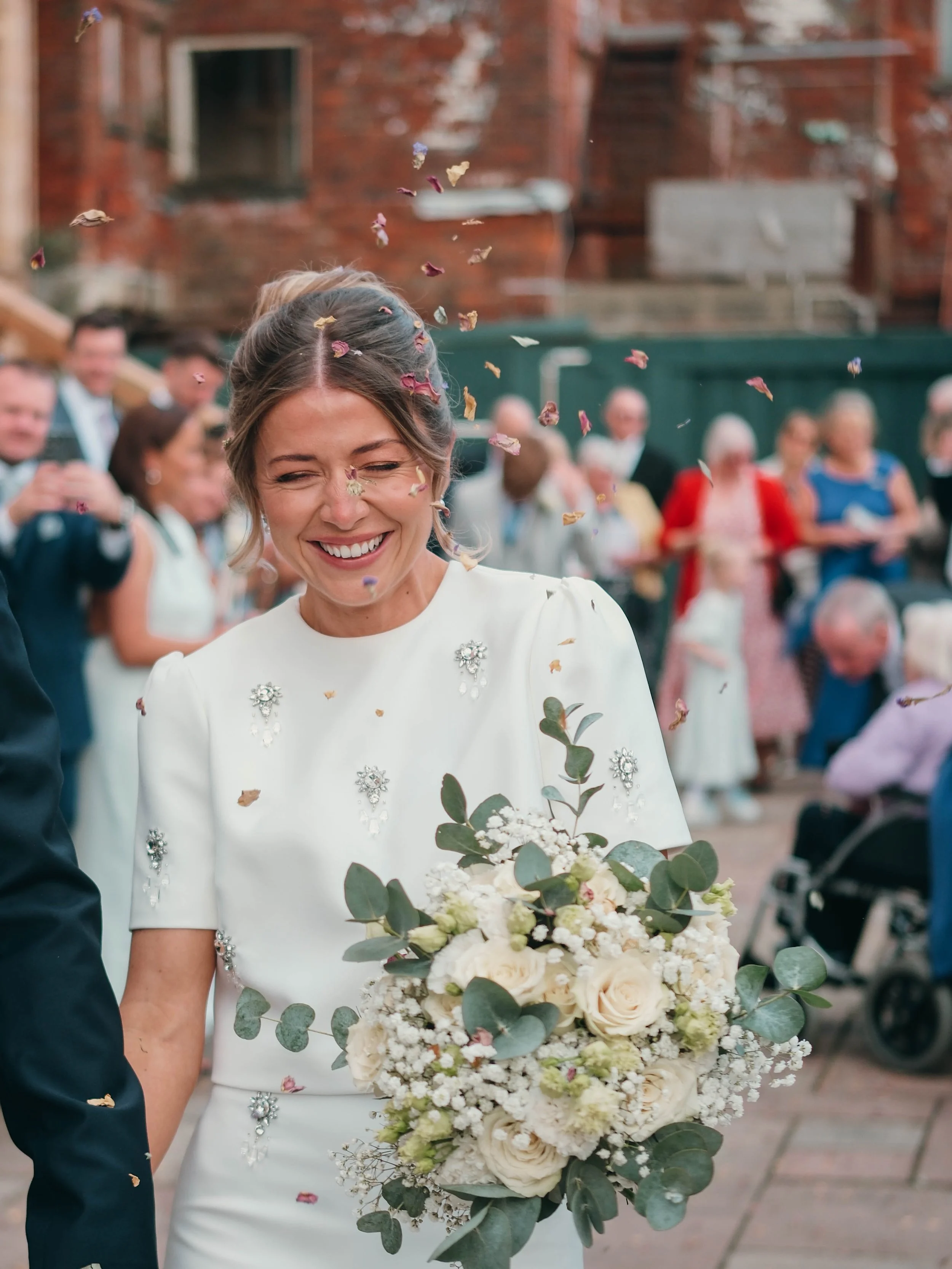 A bride holding a large bouquet of white and cream flowers, smiling as flower petals are tossed in the air during a joyful wedding celebration.