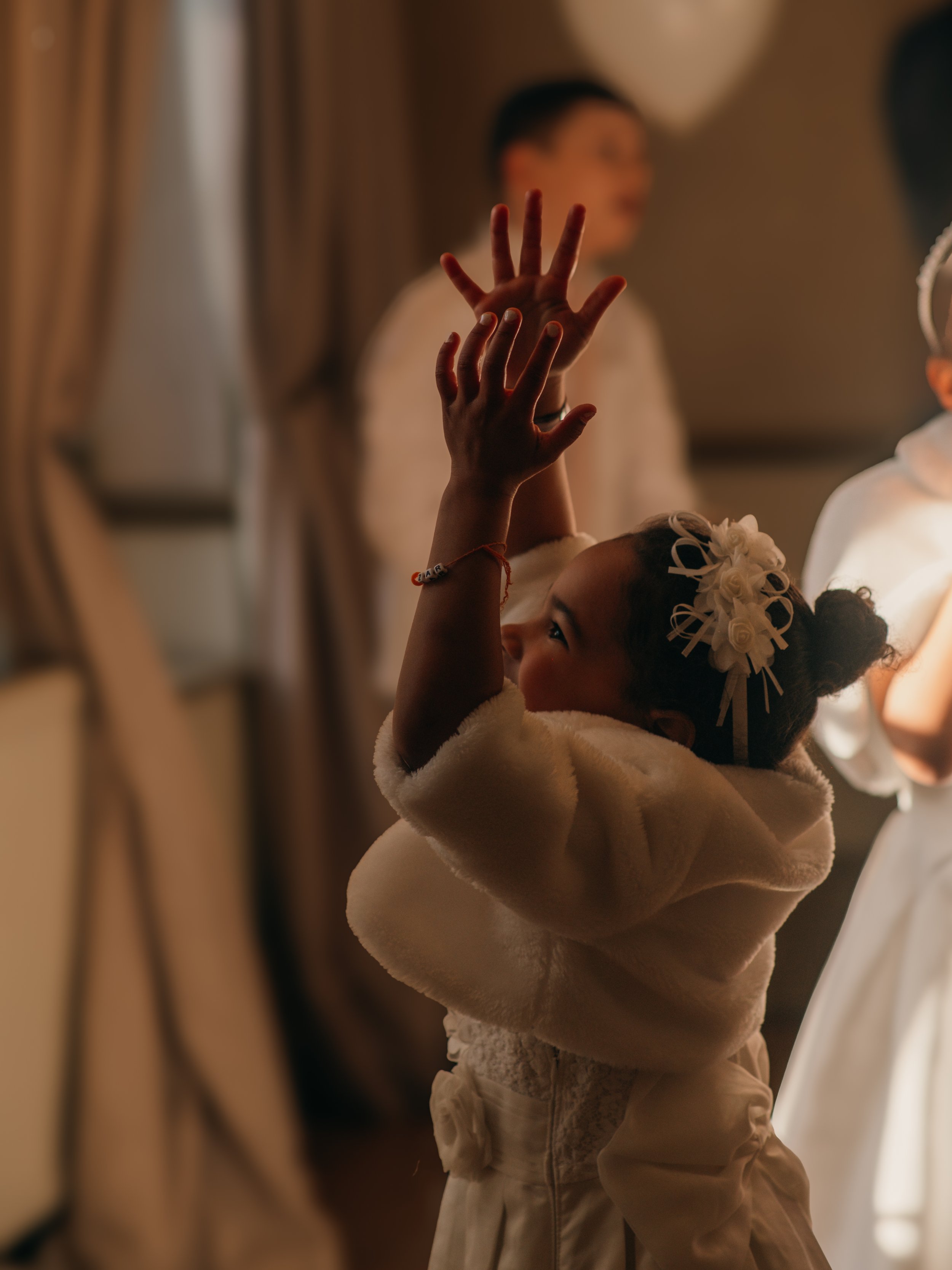 A young girl with curly hair holding her hands up to her face, wearing a white fluffy coat and a decorative headband with flowers, in a warmly lit indoor setting.