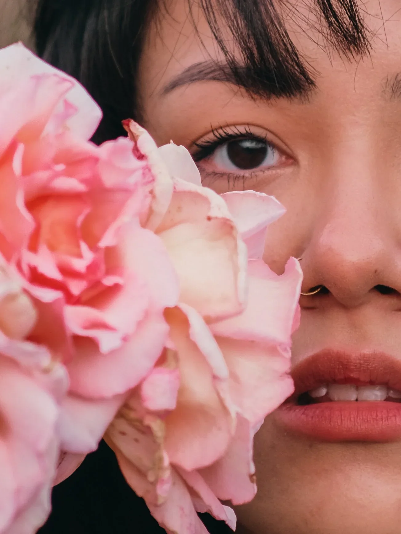 A close-up of a woman with dark hair, light skin, and a nose ring, partially obscured by a large pink flower, with only one eye visible.