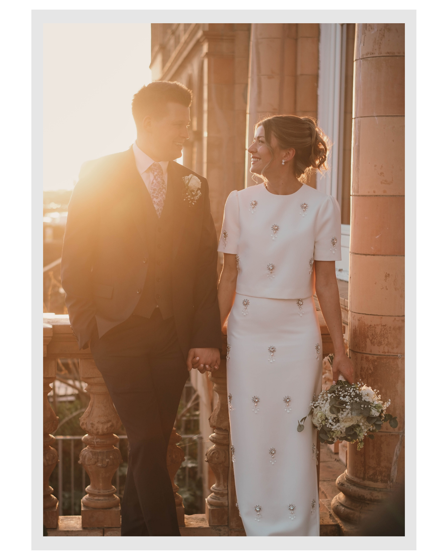 A smiling bride and groom holding hands and looking at each other outside during sunset, dressed in wedding attire, with the bride holding a bouquet of flowers.