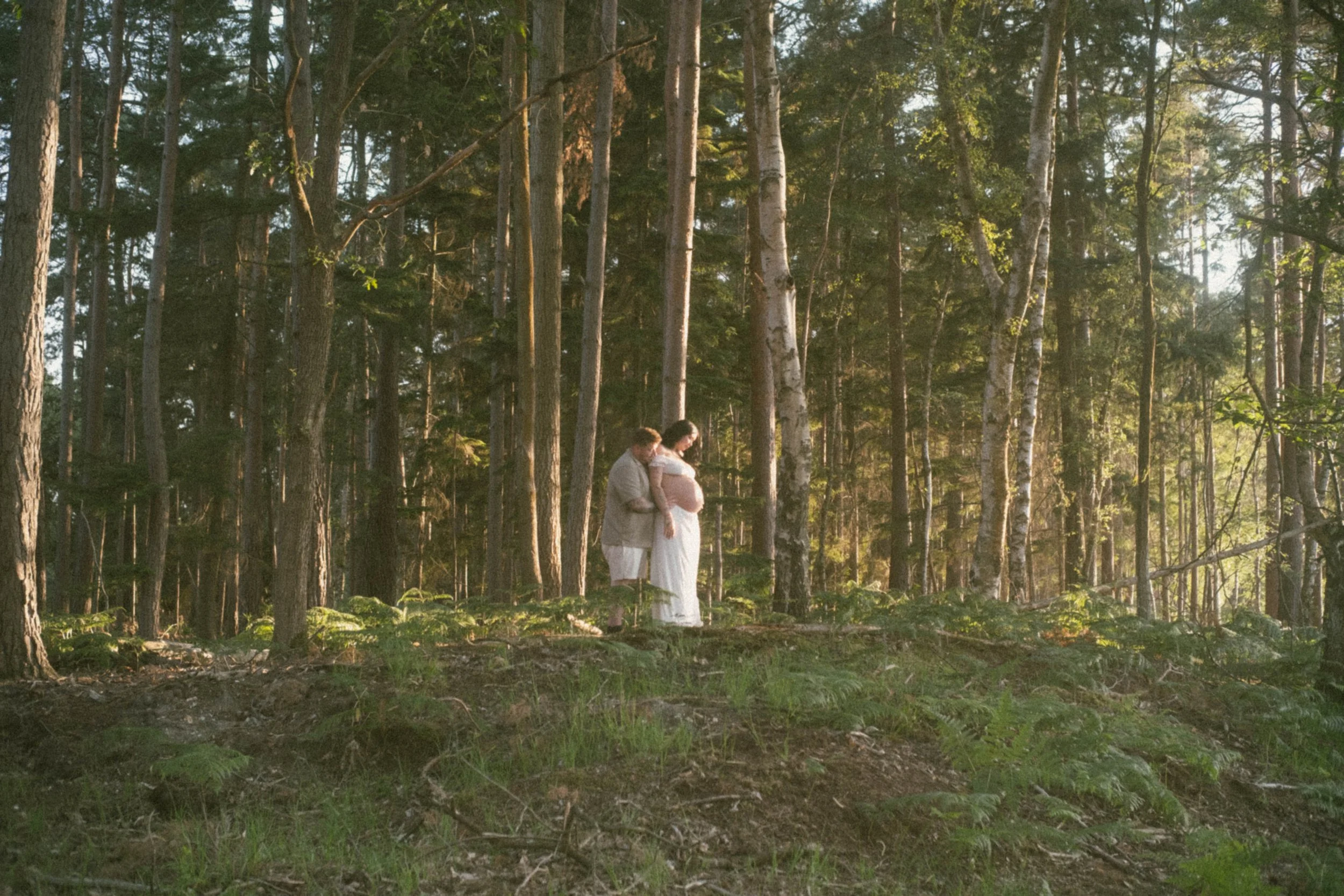 A pregnant woman and her partner standing in a forest during sunset, holding hands and looking at each other.
