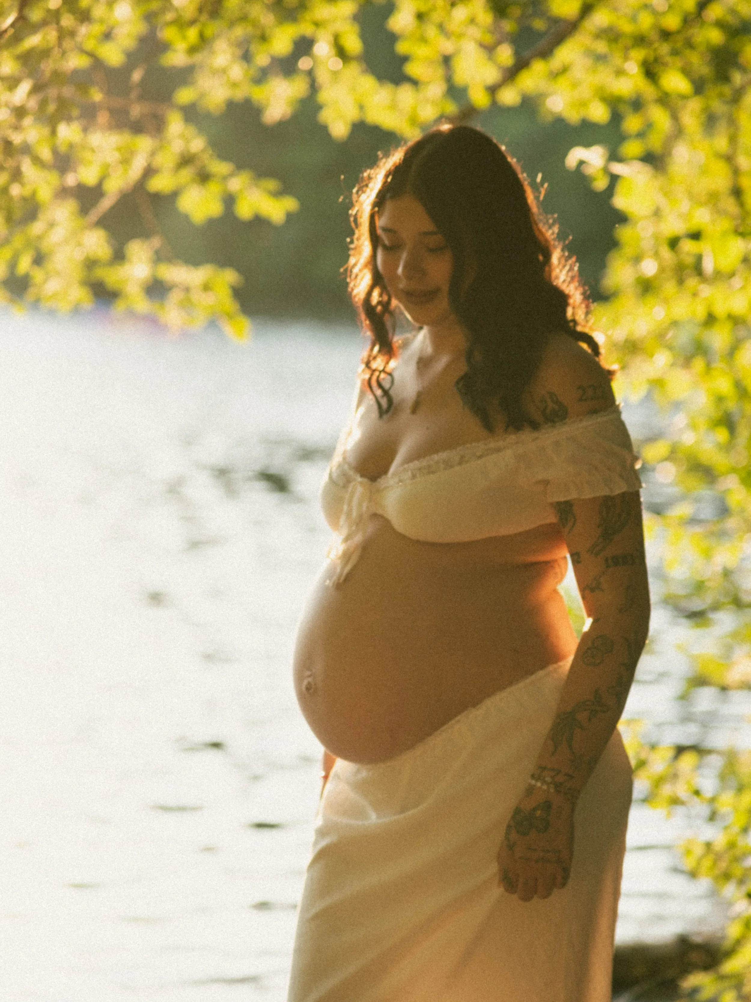 A pregnant woman standing by a body of water with trees in the background. She has tattoos on her arms and shoulder, wearing a light-colored off-shoulder top and shorts, with sunlight creating a backlit effect.