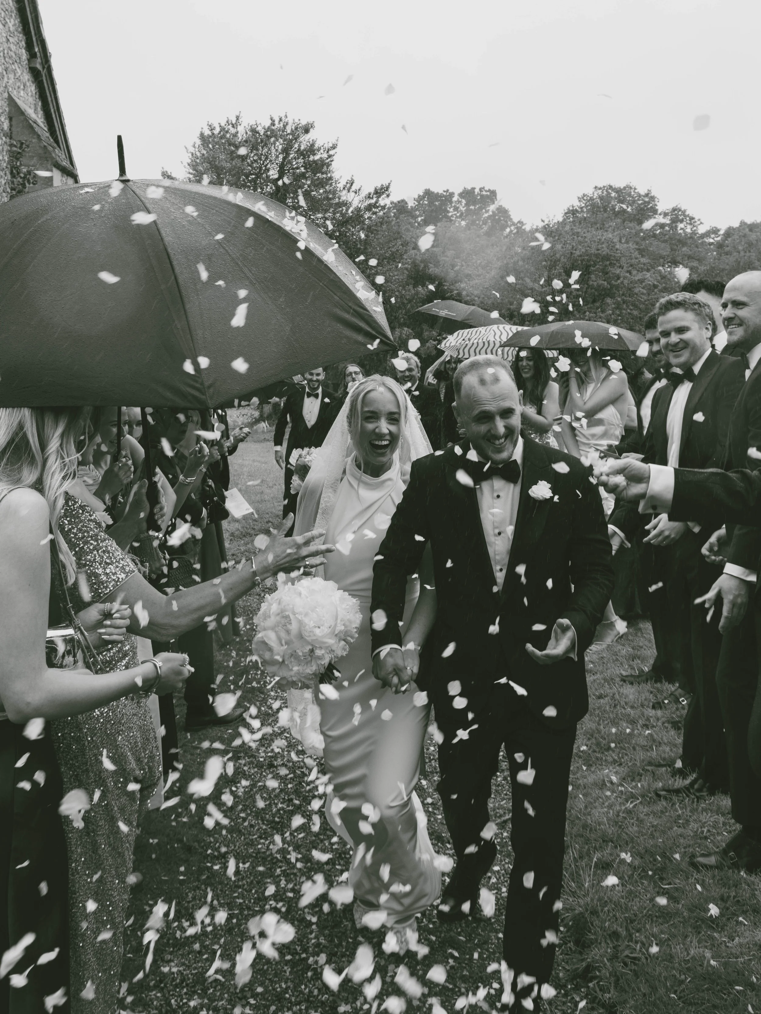 A black and white photo of a bride and groom walking down the aisle, holding hands, surrounded by guests throwing flower petals, with umbrellas overhead on a rainy day.