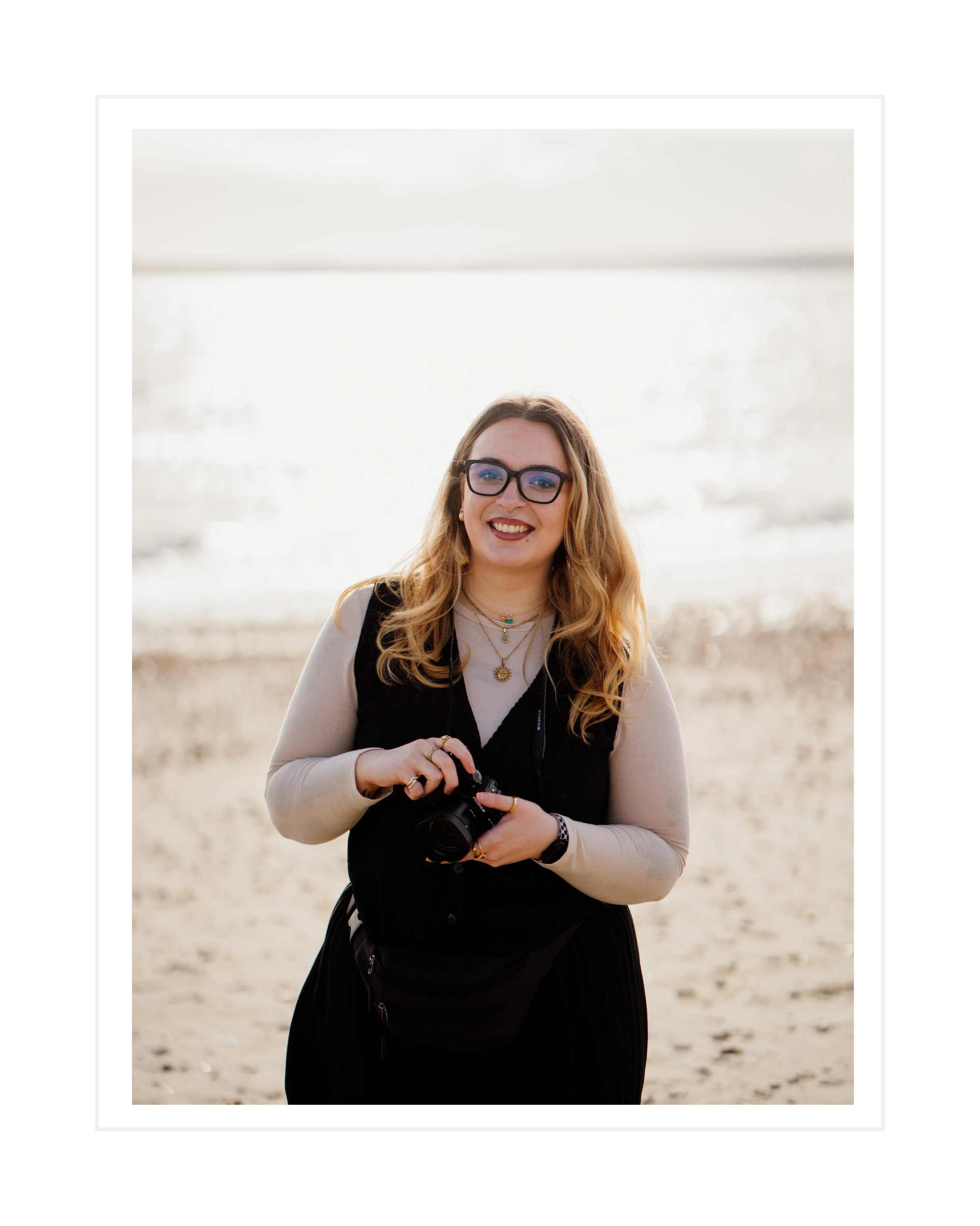 A young woman with glasses, long wavy hair, wearing a beige long-sleeve top and black vest, smiling and holding a camera, standing on a sandy beach with the ocean in the background.