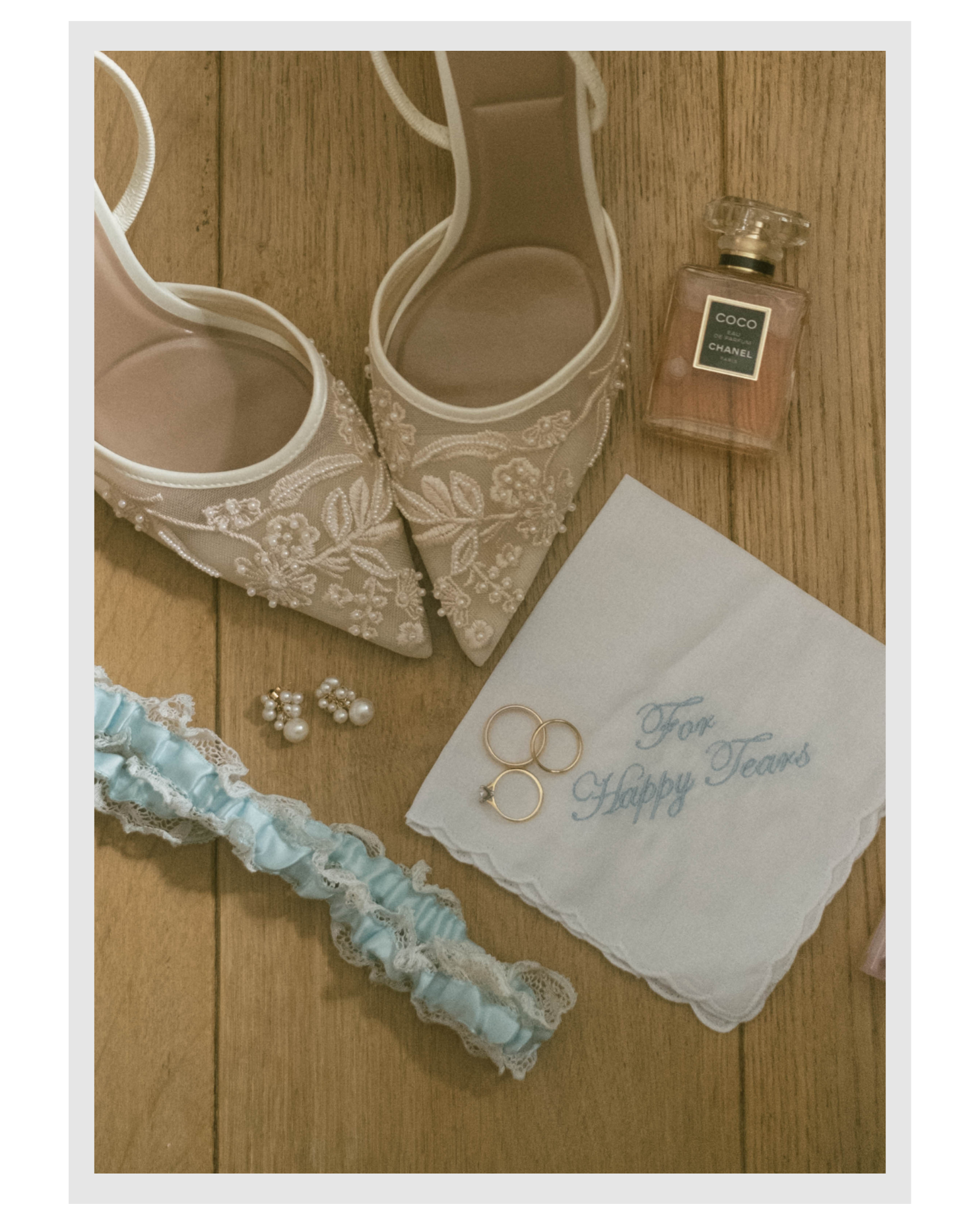 A pair of white, lace and bead embroidered bridal shoes with open toes, a light blue and white garter, pearl earrings, three gold rings on a white handkerchief with blue script, and a bottle of Chanel Coco Mademoiselle perfume on a wooden surface.