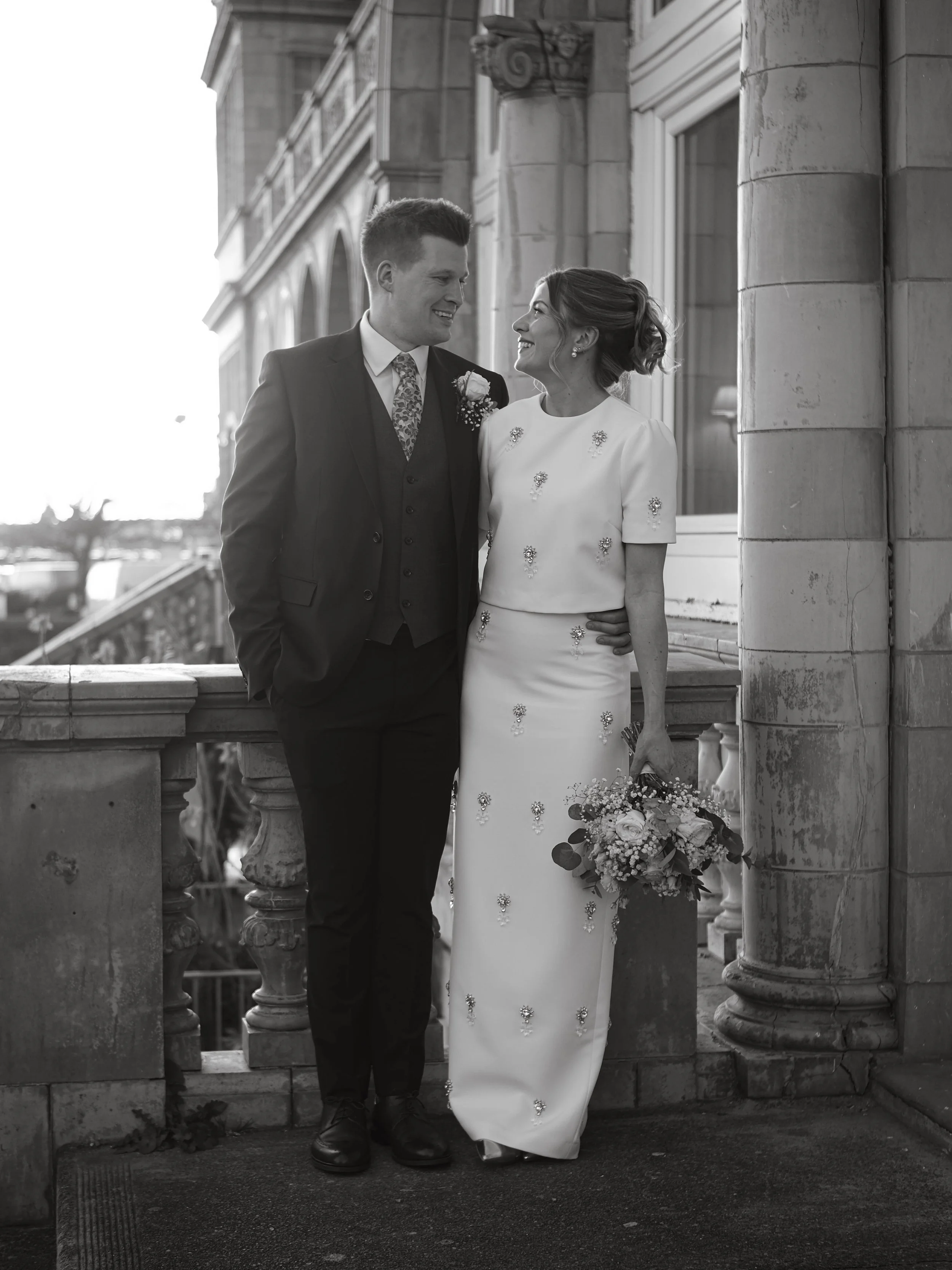 A bride and groom smiling at each other on a balcony with stone columns, dressed in wedding attire, with the bride holding a bouquet of flowers.