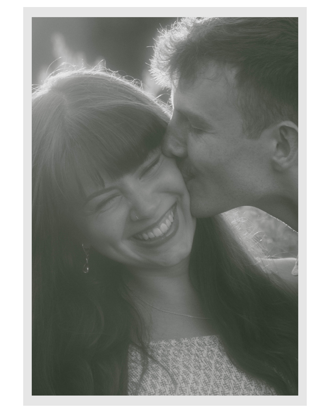 A black and white close-up of a young couple sharing a joyful moment, with the man kissing the woman's cheek and both smiling brightly.
