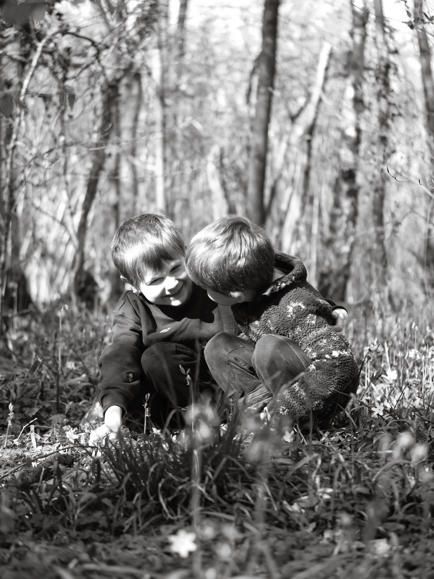 Two young boys sitting closely together in a wooded area, smiling and enjoying each other's company on a day outdoors.