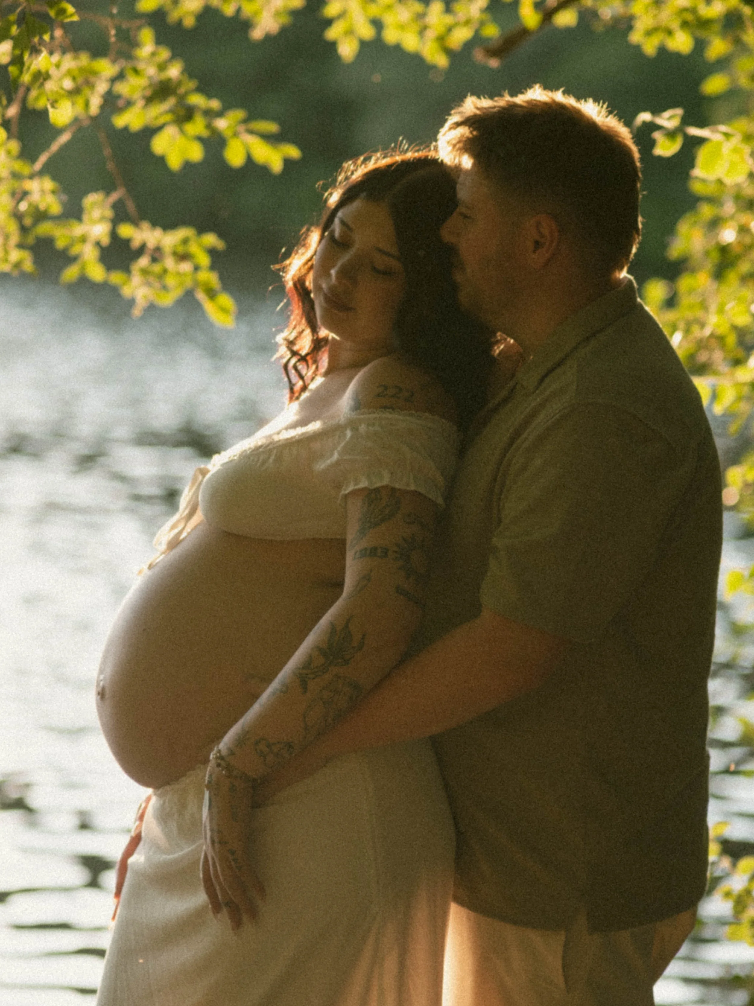 A couple embracing outdoors near a body of water with trees and green foliage in the background.