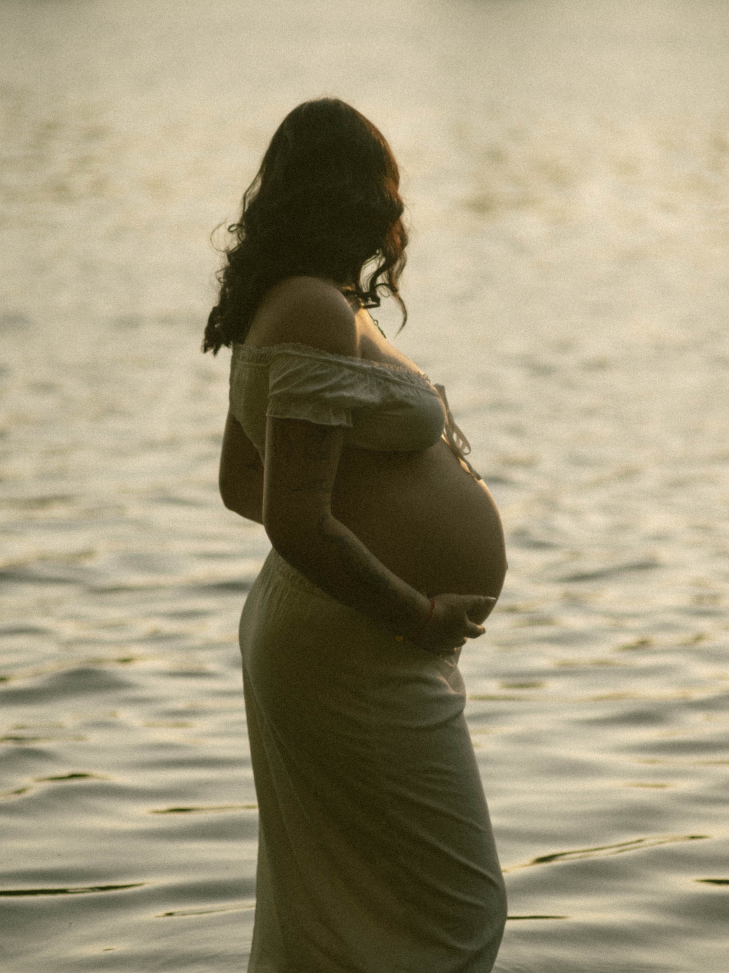 A pregnant woman standing by water during sunset, cradling her belly with both hands.