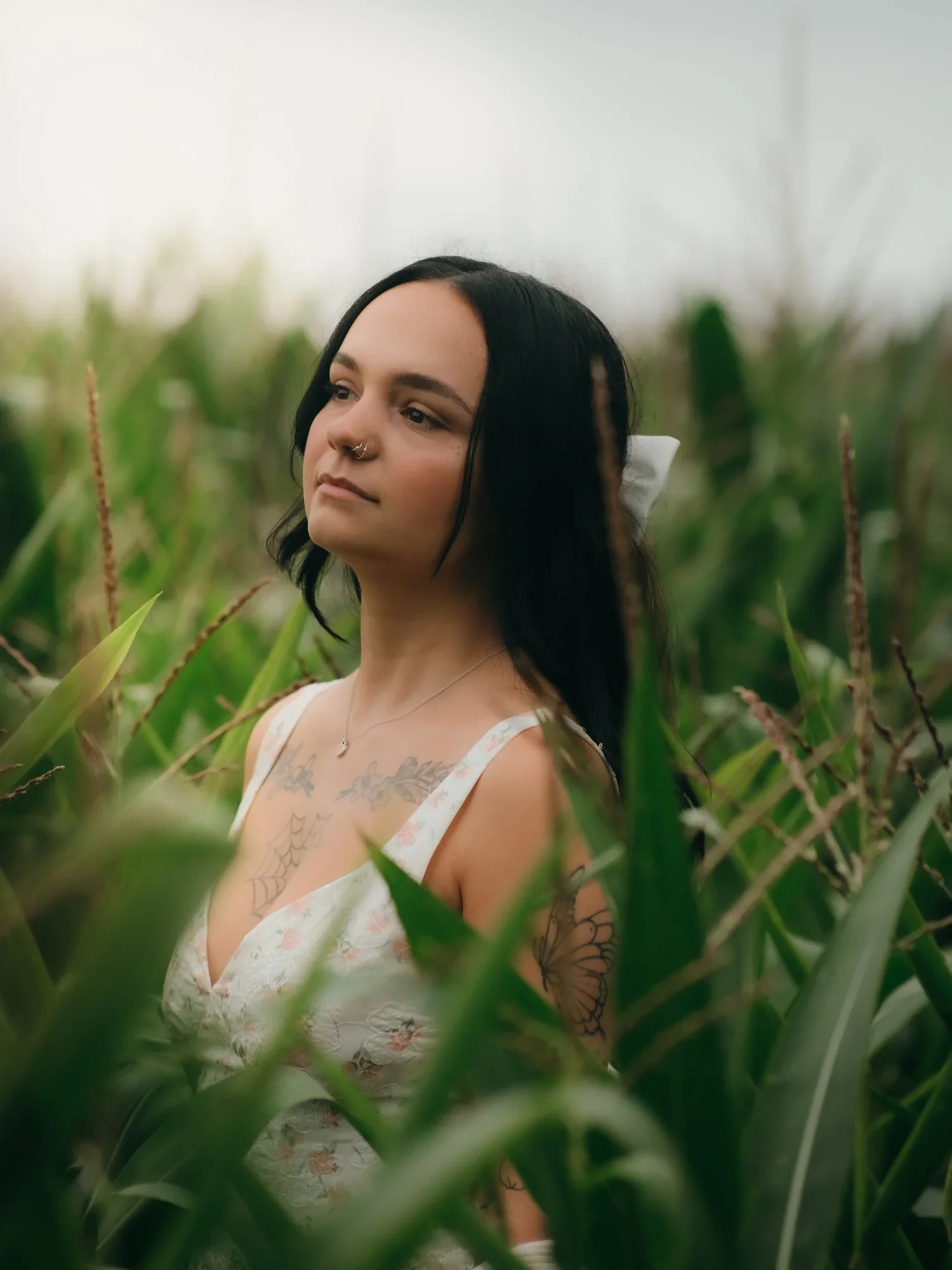 A young woman with black hair, tattoos on her arm and chest, wearing a sleeveless floral dress, standing amidst green corn plants outdoors.