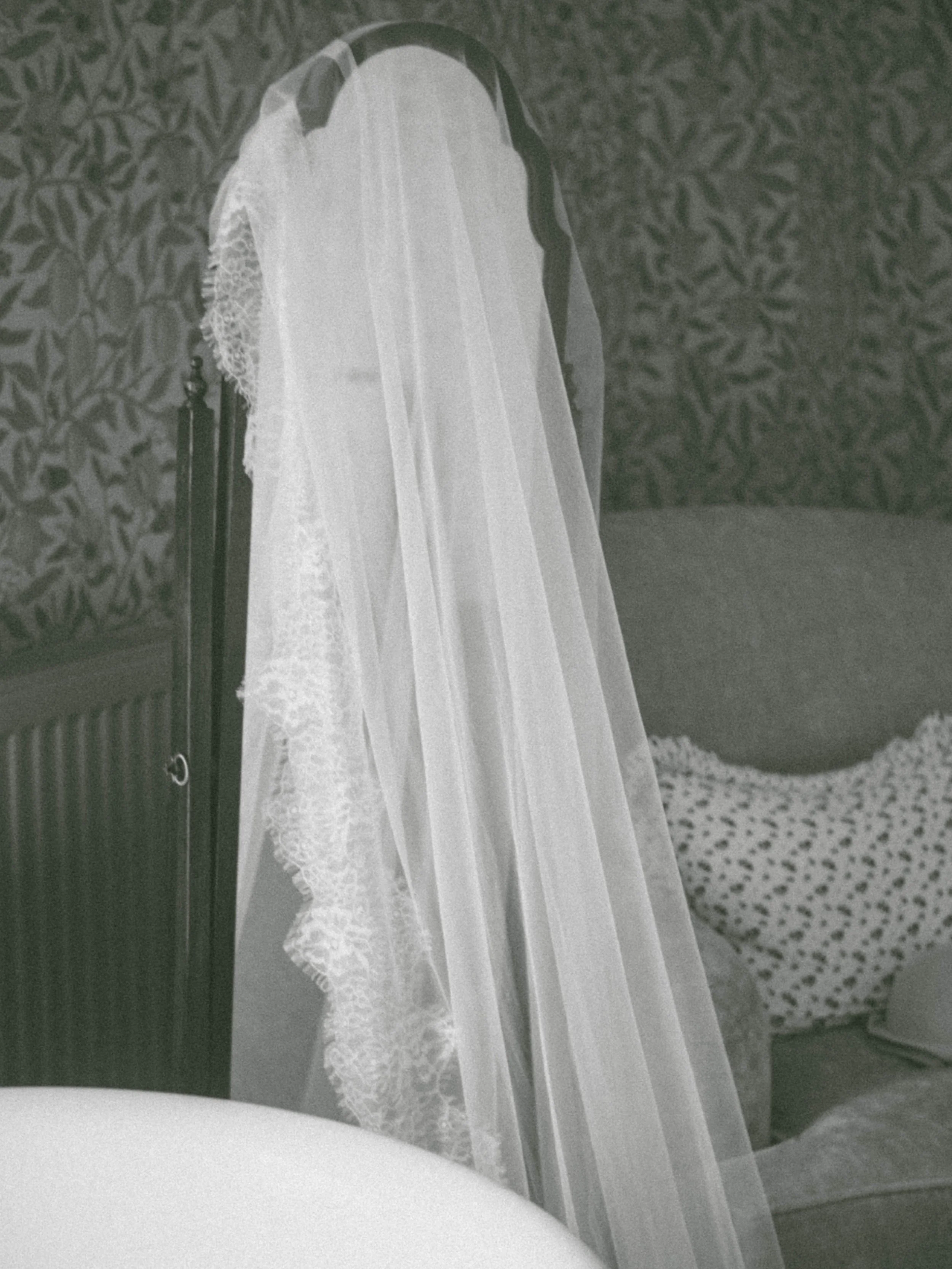 A black and white photo of a wedding veil hanging on a mirror, with a vanity and a patterned bedspread in the background.