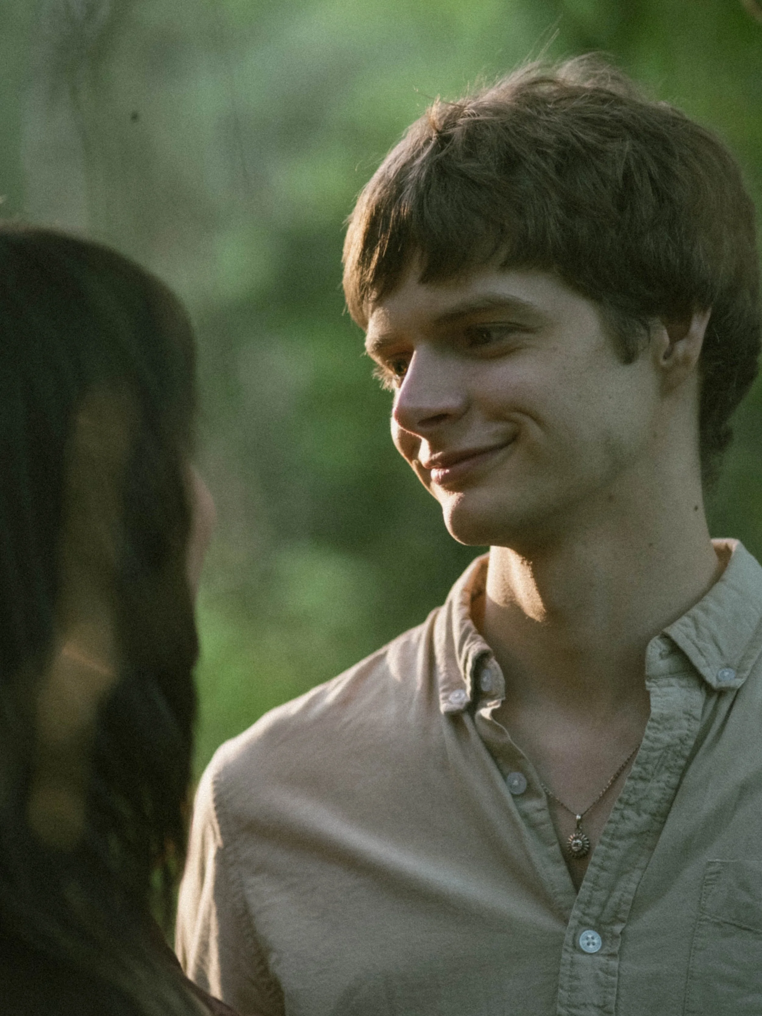 A young man with short brown hair smiling and looking at a woman with long dark hair, both outdoors with a blurred green background.