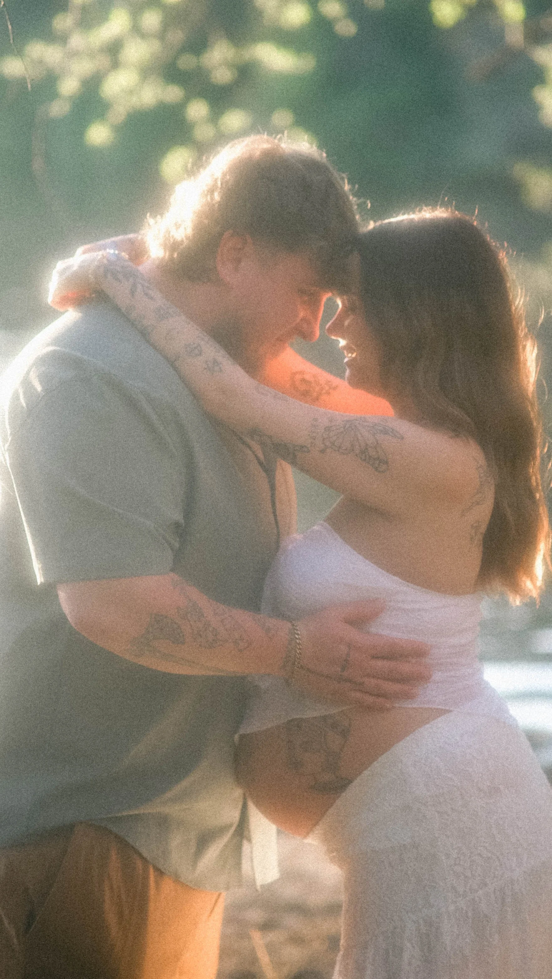 A young couple embracing each other, smiling and looking into each other's eyes outdoors during sunset, with blurred green foliage in the background.