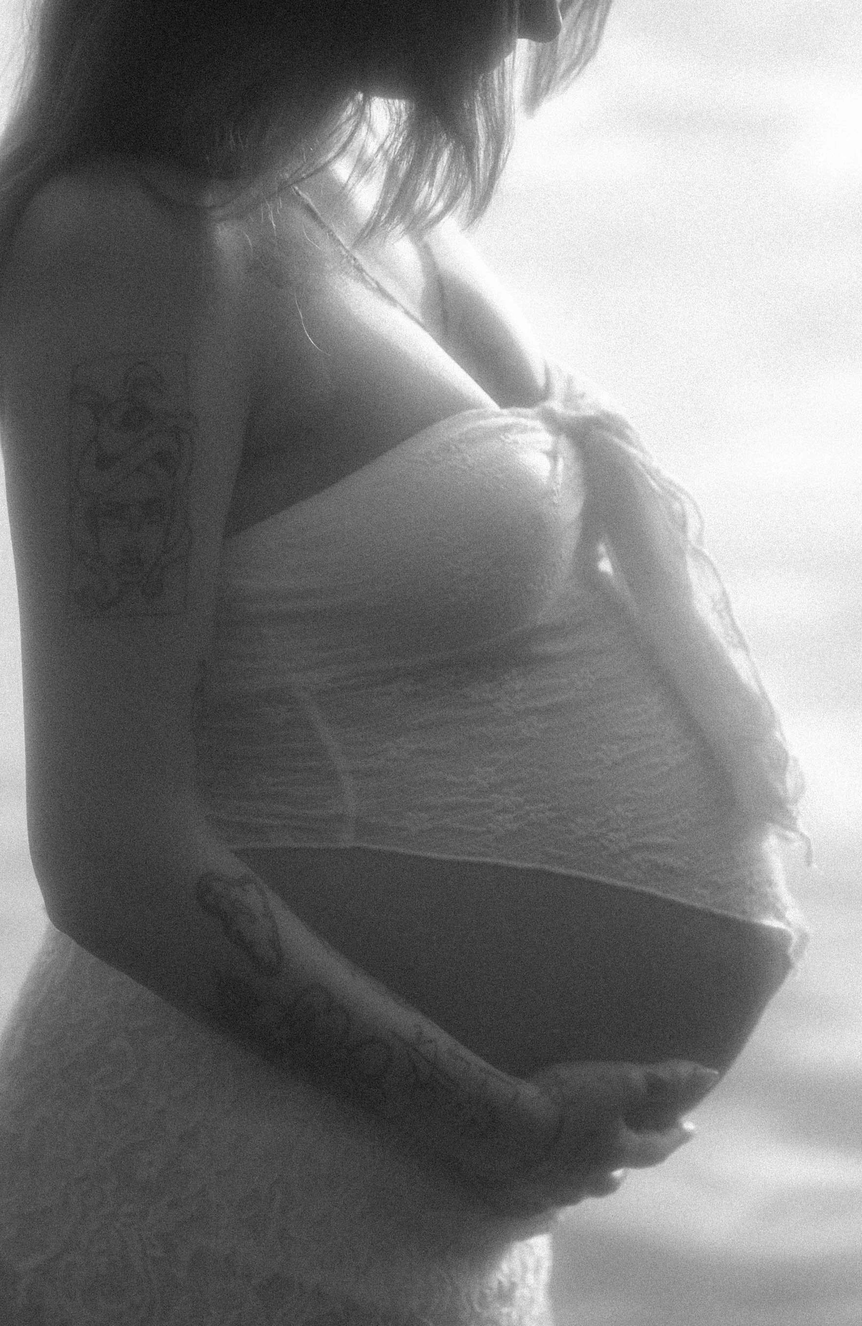 Close-up black and white photo of a woman with curly hair and tattoos, wearing a strapless dress, sitting and looking down.