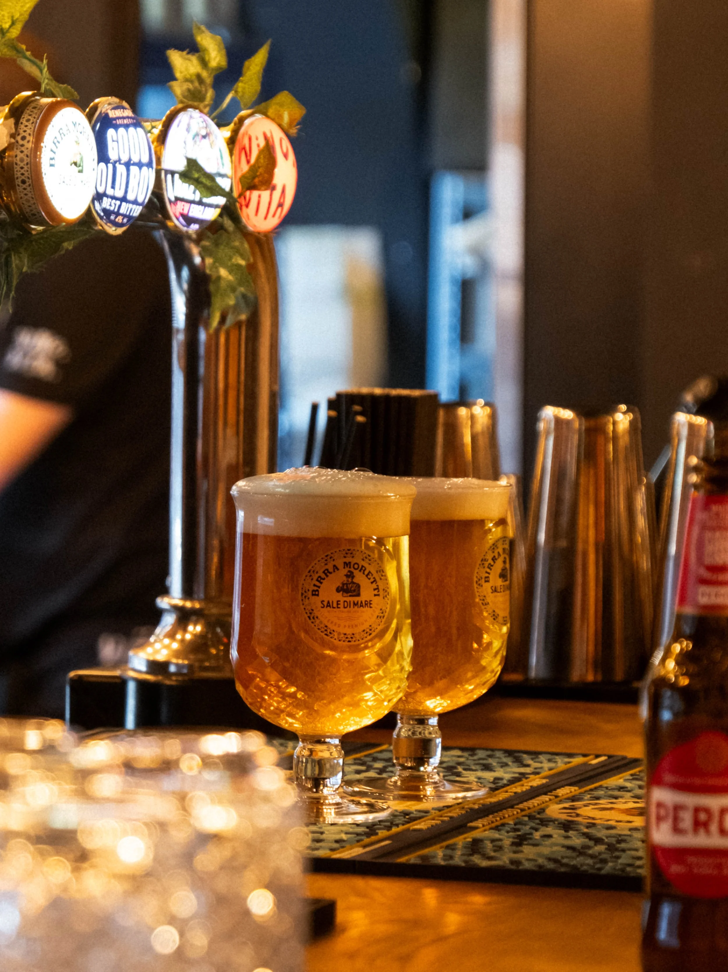 Three glasses of beer on a bar counter with bar taps in the background, illuminated by warm lighting.