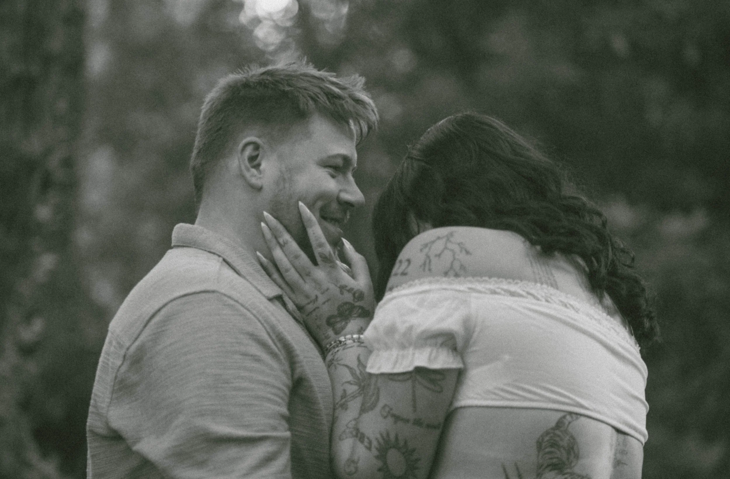 A grayscale photo of a young man and woman closely interacting outdoors. The woman is touching the man's face and they are smiling with an emotional connection.