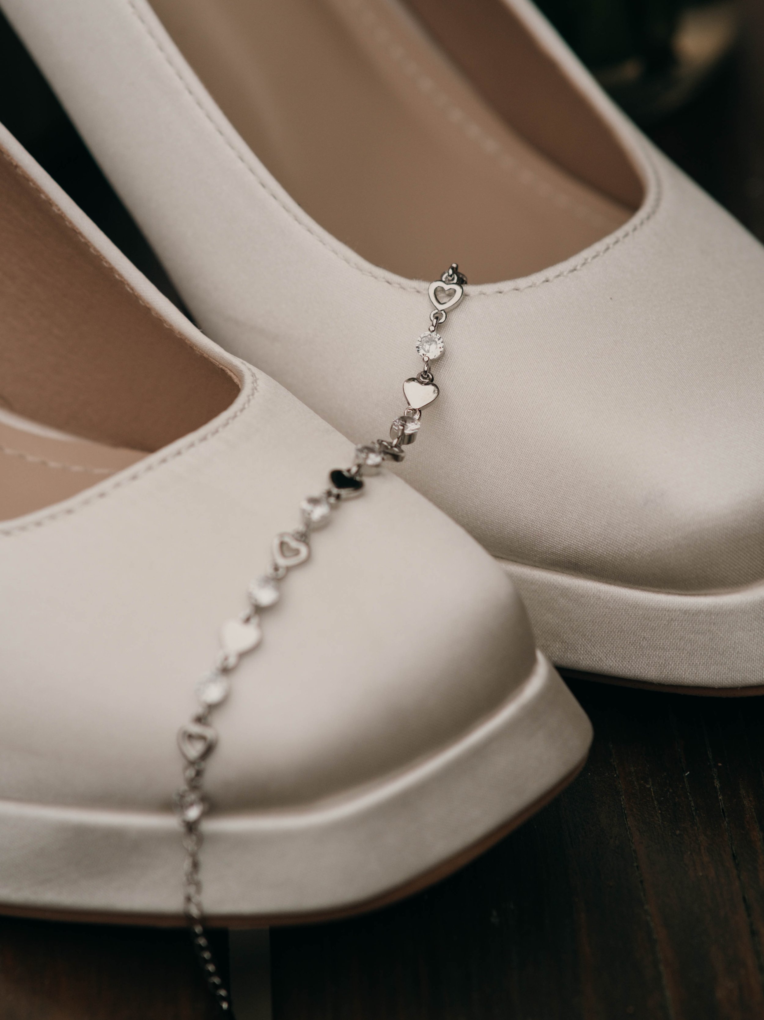 Close-up of a silver charm bracelet with hearts and a crystal, resting on a cream-colored shoe and a white box.