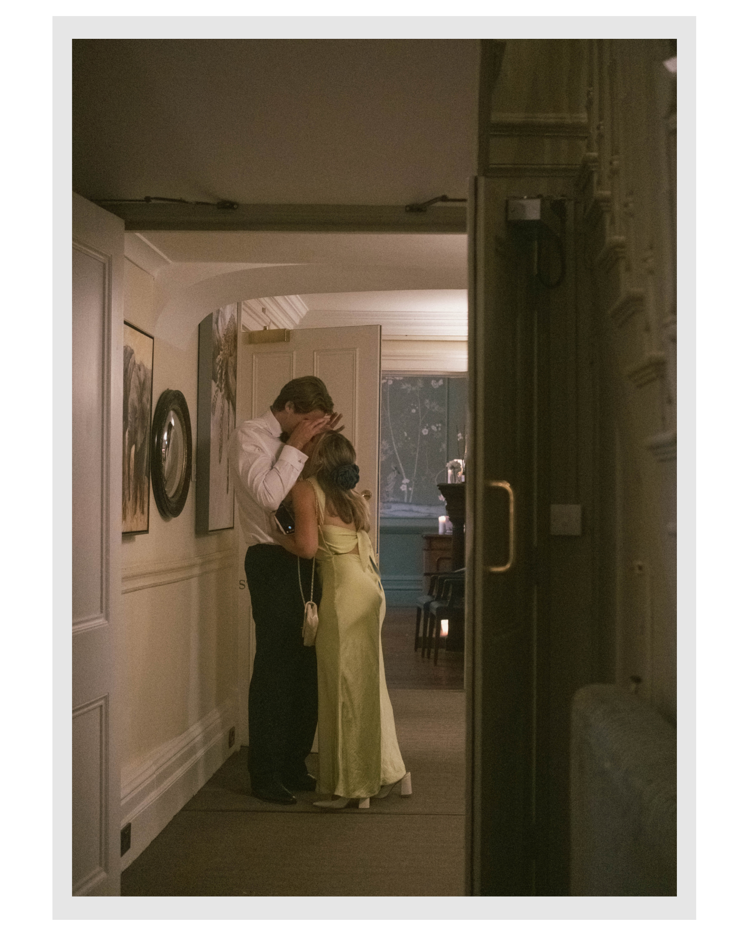 Couple dressed in formal attire, standing together indoors under warm lighting, with the man holding the woman's hands and the woman wearing a long yellow gown.