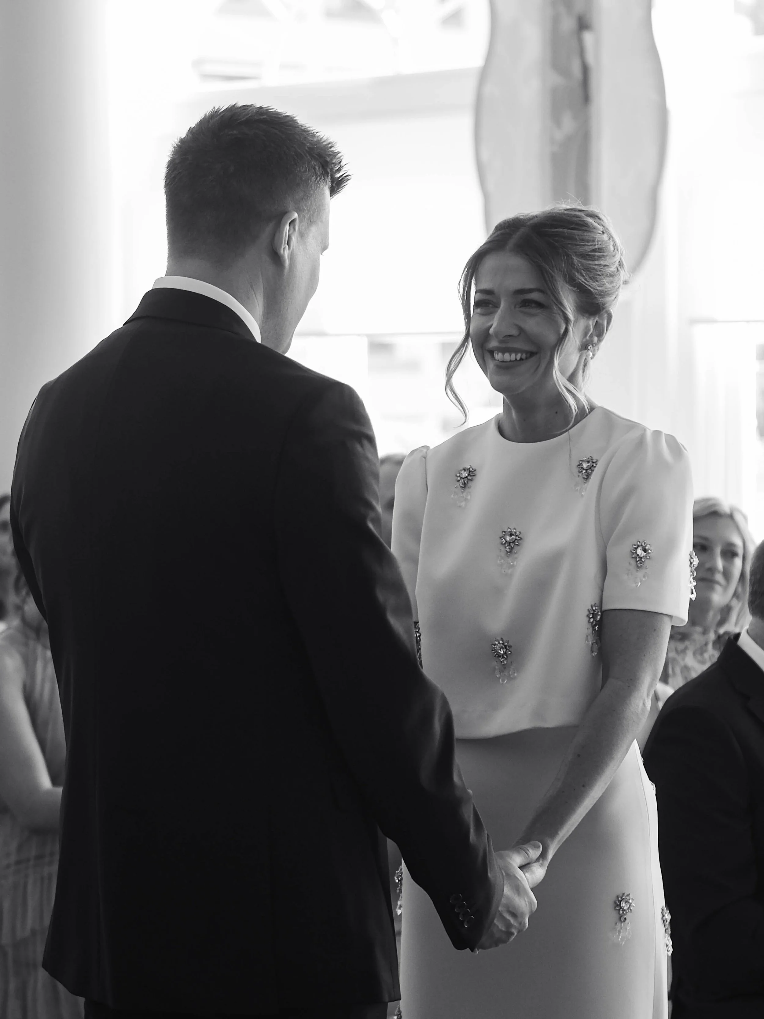A black and white photo of a wedding ceremony showing a groom and bride holding hands, with the groom's back to the camera and the bride smiling at him, in front of a cross.
