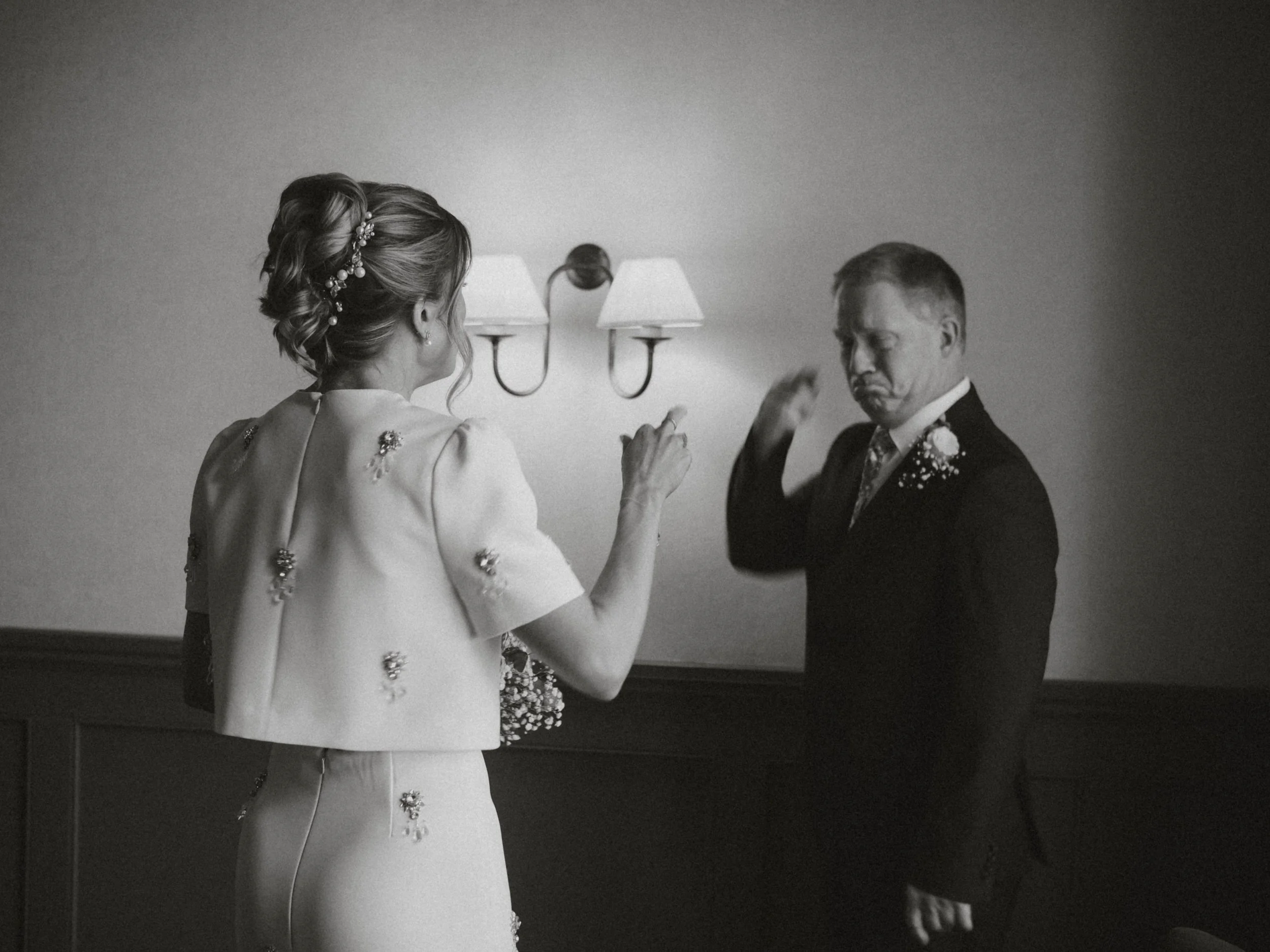 A bride with an elegant hairstyle and floral hair accessories wearing a wedding dress, standing in front of a groom in a tuxedo with a boutonniere, in a dimly lit indoor setting.