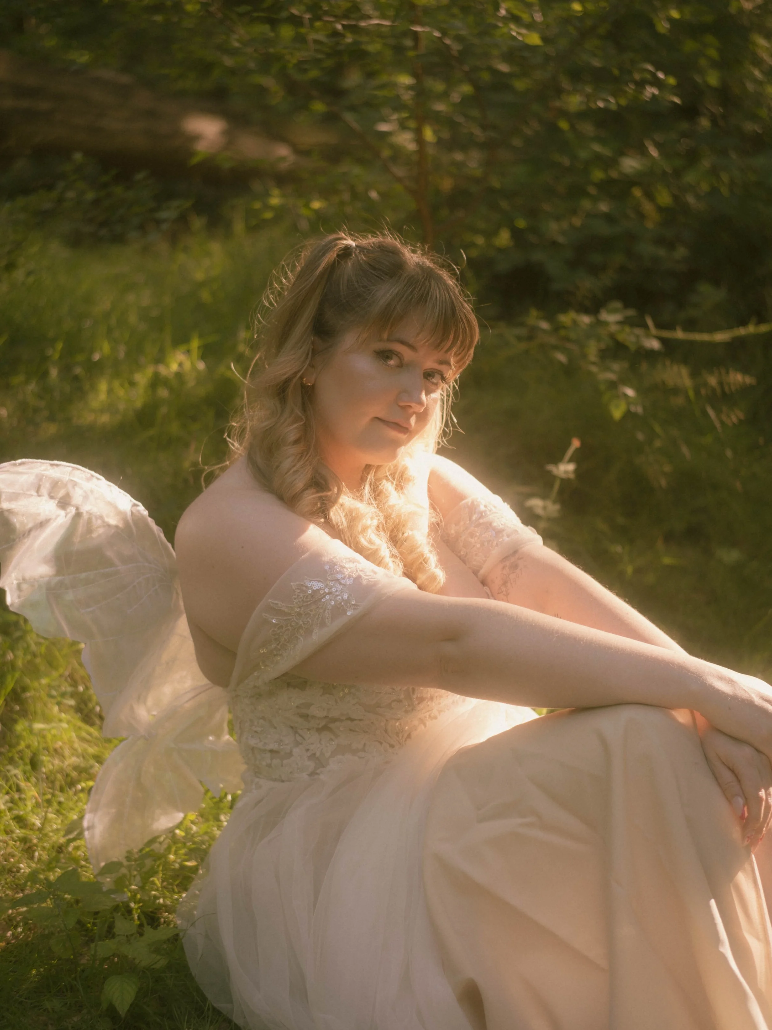 A woman with wavy blonde hair wearing a white lace dress, sitting outdoors in a grassy area with green foliage in the background, bathed in warm sunlight.