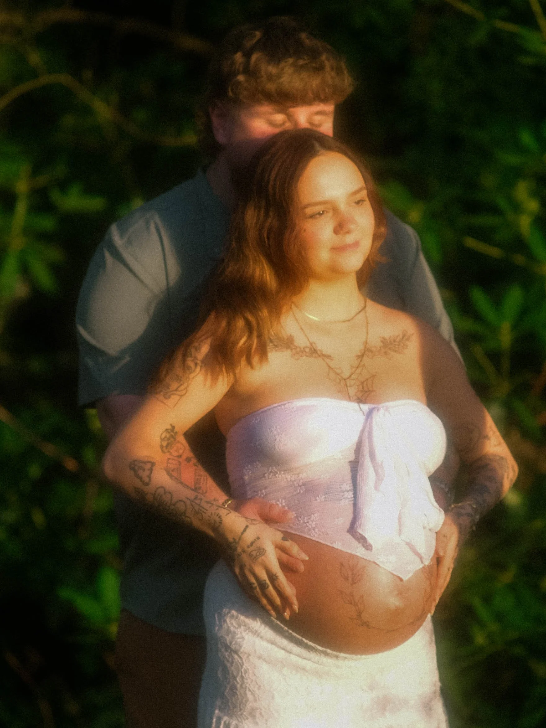 A woman with reddish-brown wavy hair, tattoos on her arms and chest, wearing a strapless white lace top, standing outdoors at night with a person behind her, surrounded by green bushes.