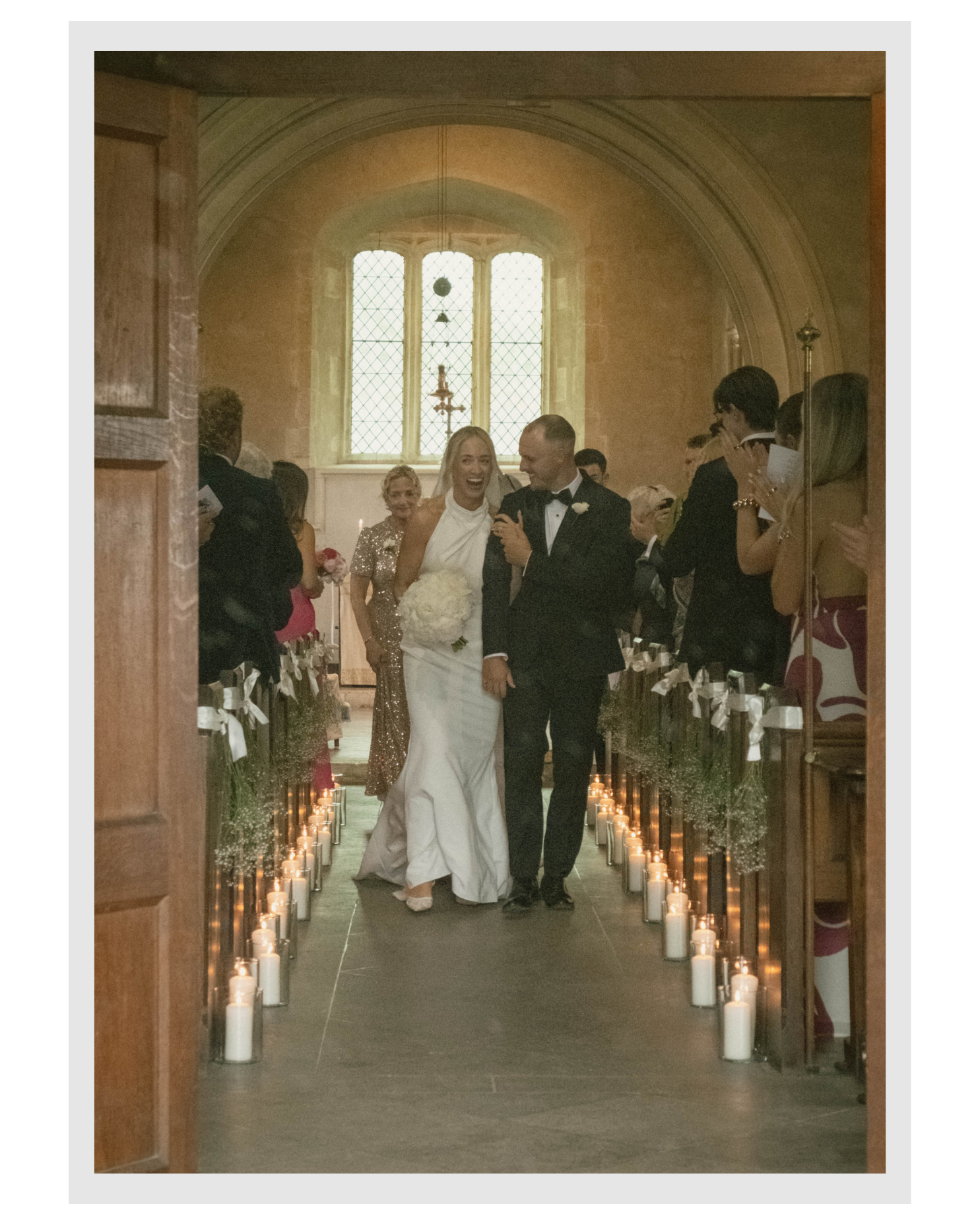 A bride and groom walking down the aisle of a church, smiling and laughing, surrounded by friends and family, with candles lining the aisle, during a wedding ceremony.