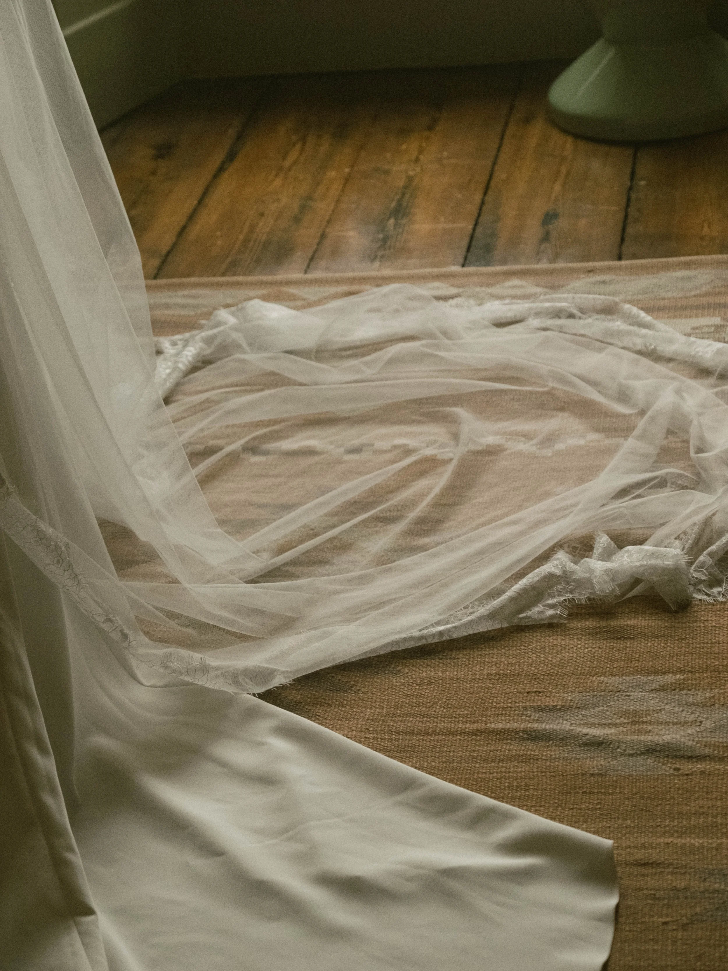 A sheer white curtain is draped over a bed with a beige-striped bedspread, with a wooden floor visible in the background.