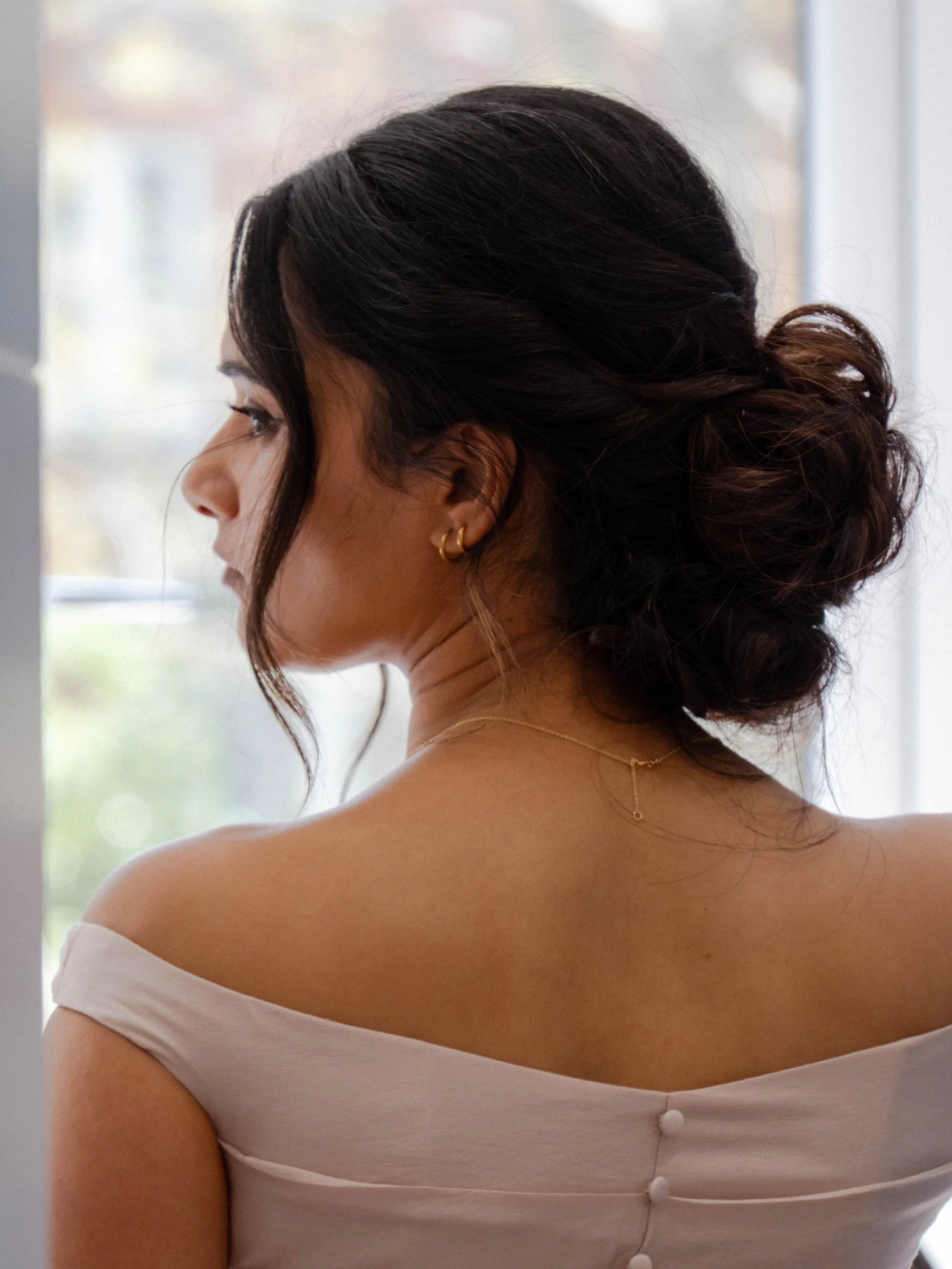 Side view of a woman with dark brown hair styled in an elegant updo, wearing gold hoop earrings and a delicate necklace, dressed in an off-the-shoulder white top with decorative buttons, near a window with soft natural light.