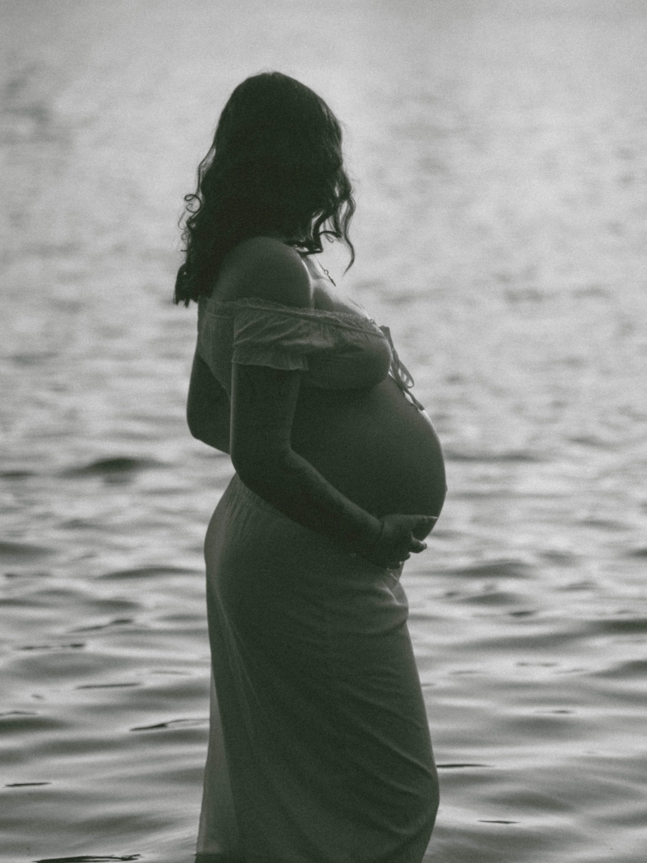 A pregnant woman with long, wavy hair stands in the ocean, facing sideways with her hands on her belly.