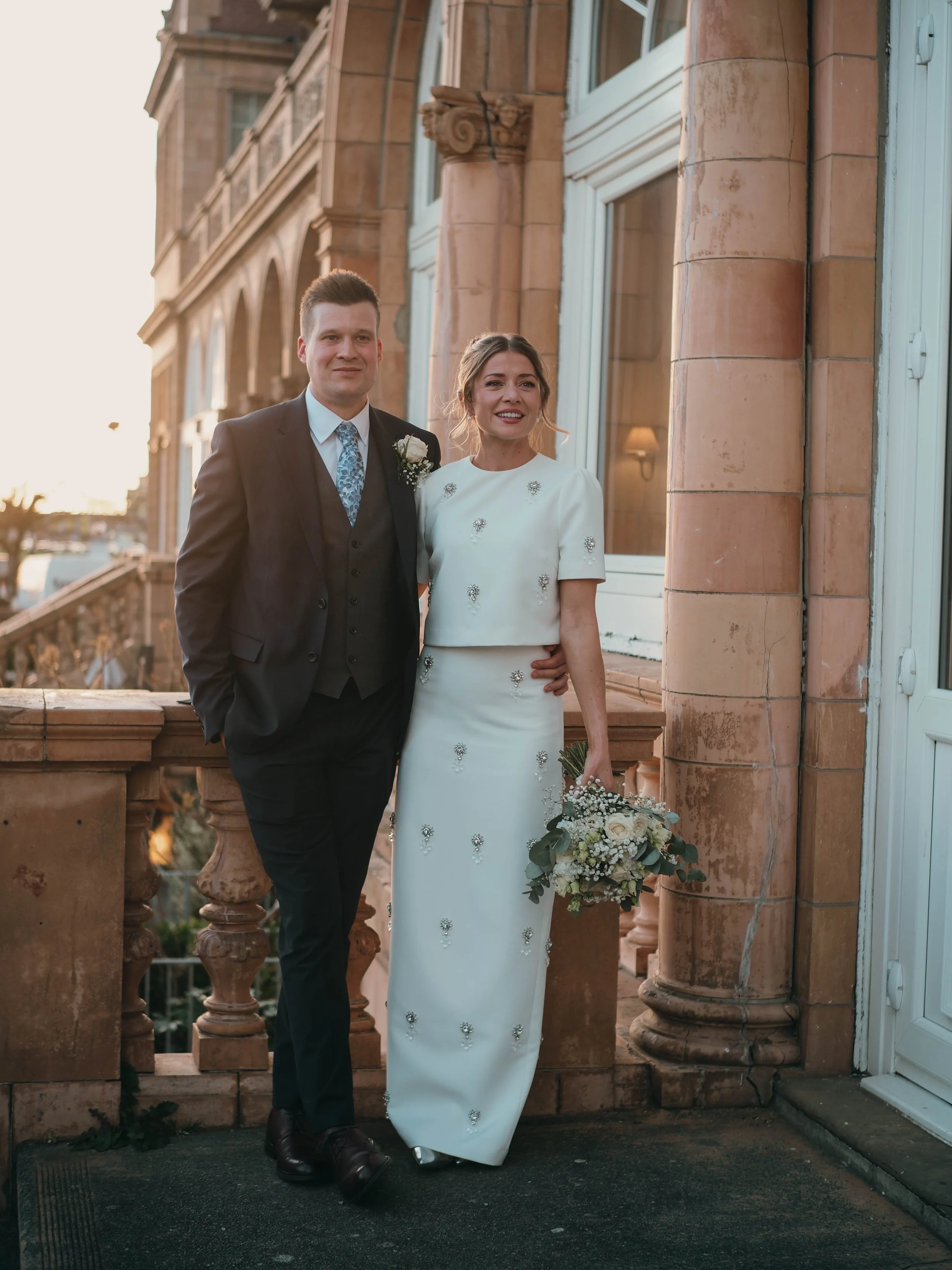 A newlywed couple standing on a balcony in front of a building with stone columns. The man is dressed in a dark suit with a patterned tie and a white boutonniere. The woman is wearing a white wedding dress with embellishments, holding a bouquet, and 