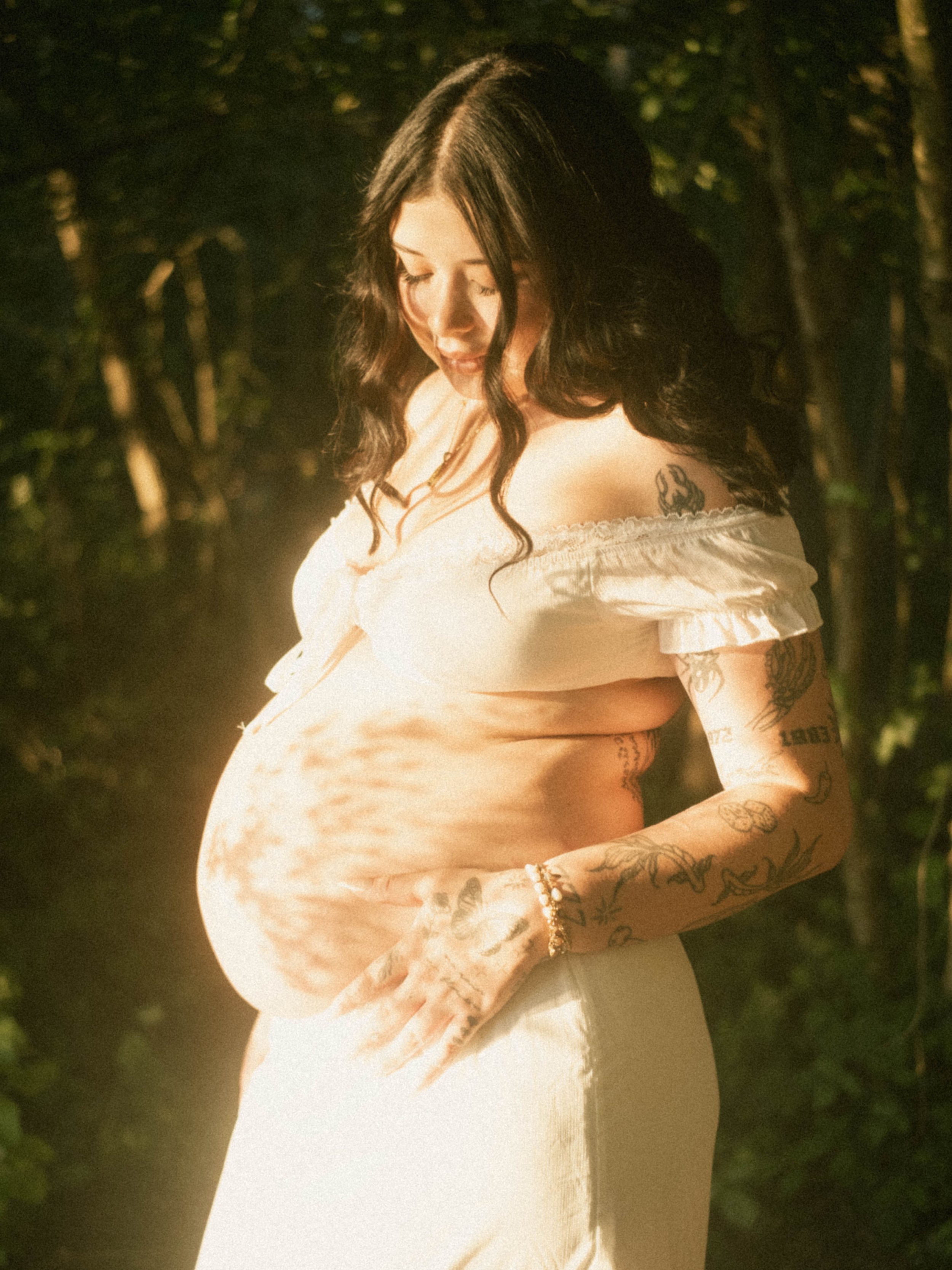A pregnant woman with dark hair and tattoos on her arms and shoulder, wearing a light-colored off-shoulder top, standing outdoors surrounded by trees, with sunlight illuminating her face and belly.