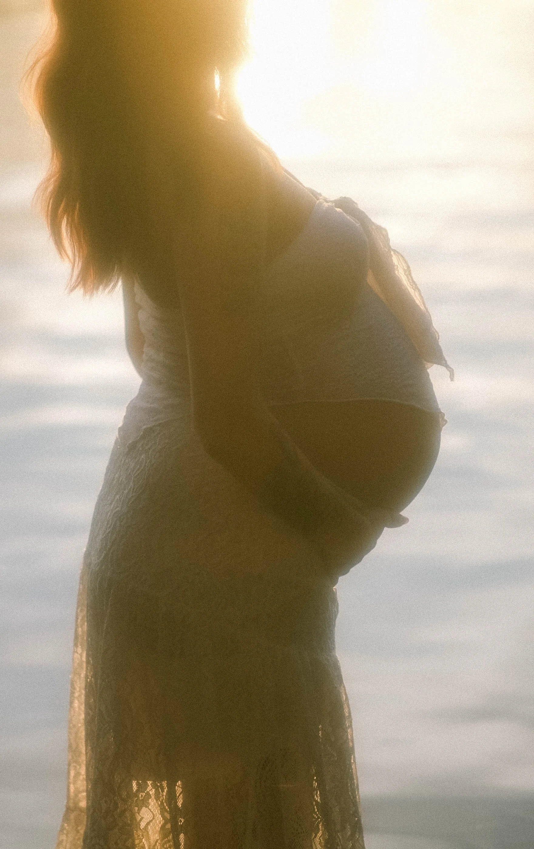Silhouette of a woman with long hair, wearing a dress, standing against a bright sky or water background, backlit by sunlight.