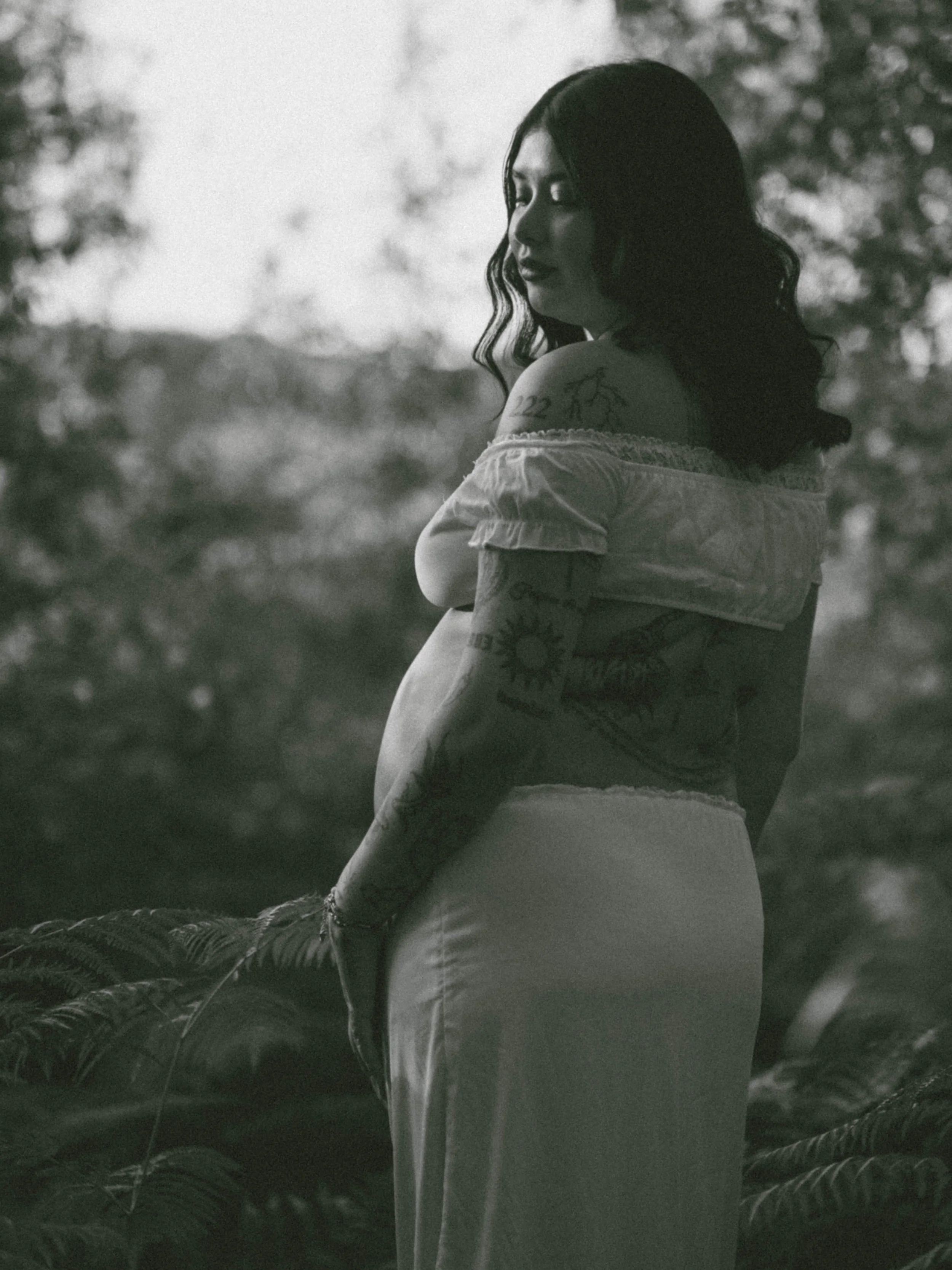 A woman with long dark hair in a loose wave, wearing an off-the-shoulder top, standing outdoors among trees, looking to the side with a thoughtful expression. She has tattoos on her shoulder and arm, and the photo appears to be taken in black and whi