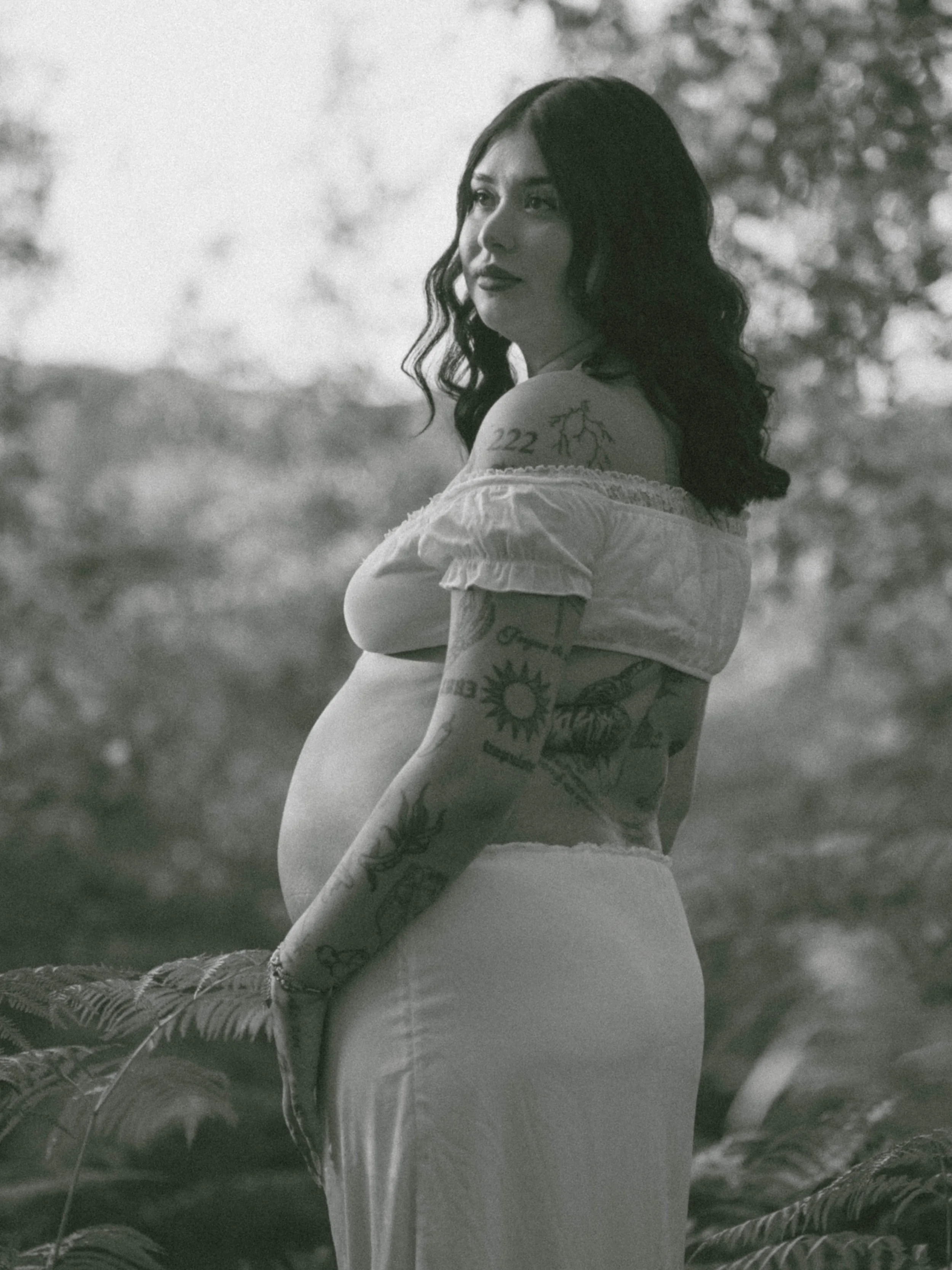 A young woman with dark, curly hair and tattoos on her shoulder and arm, wearing an off-the-shoulder top, sits outdoors in a natural setting with trees and sunlight in the background.
